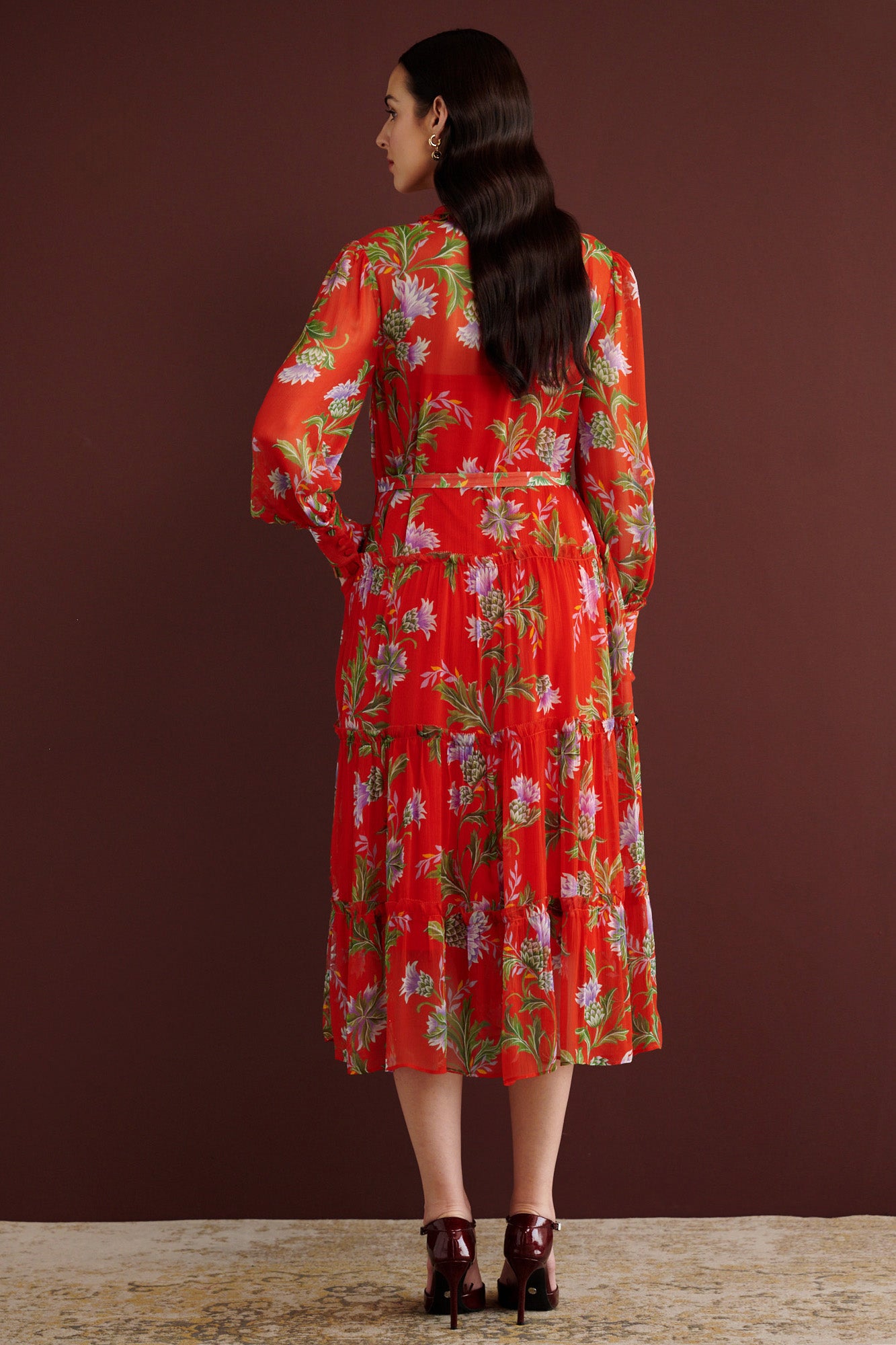 Back view of woman standing. She is wearing the Emira dress in the Stuart Thistle print. It is a flowing Flame-colored crinkle chiffon dress with sheer sleeves, delicate ruffles, fabric-covered buttons, and a detachable belt, featuring floral motifs inspired by the Scottish Highlands.