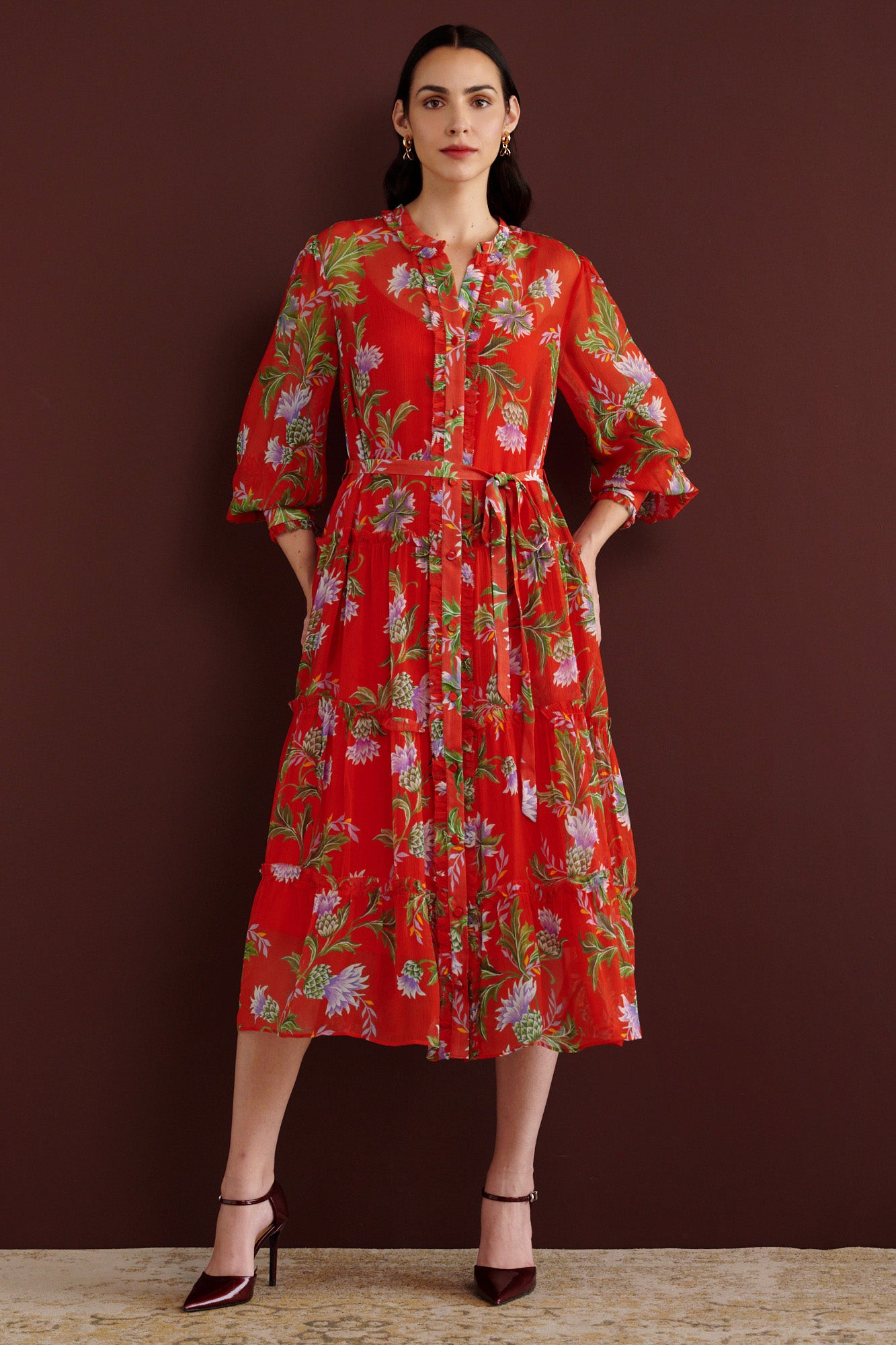 Front view of woman standing. She is wearing the Emira dress in the Stuart Thistle print. It is a flowing Flame-colored crinkle chiffon dress with sheer sleeves, delicate ruffles, fabric-covered buttons, and a detachable belt, featuring floral motifs inspired by the Scottish Highlands.