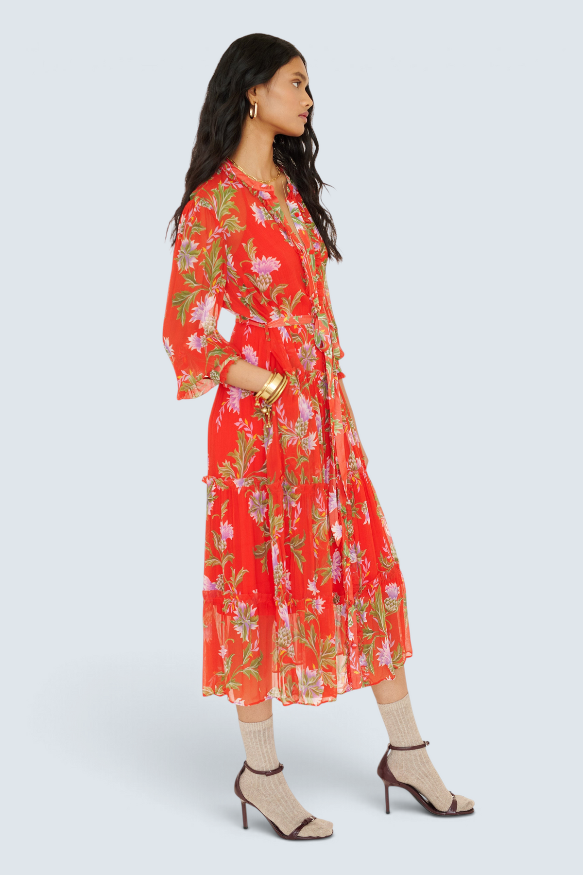 Side view of woman standing with a hand in pocket. She is wearing the Emira dress in the Stuart Thistle print. It is a flowing Flame-colored crinkle chiffon dress with sheer sleeves, delicate ruffles, fabric-covered buttons, and a detachable belt, featuring floral motifs inspired by the Scottish Highlands.
