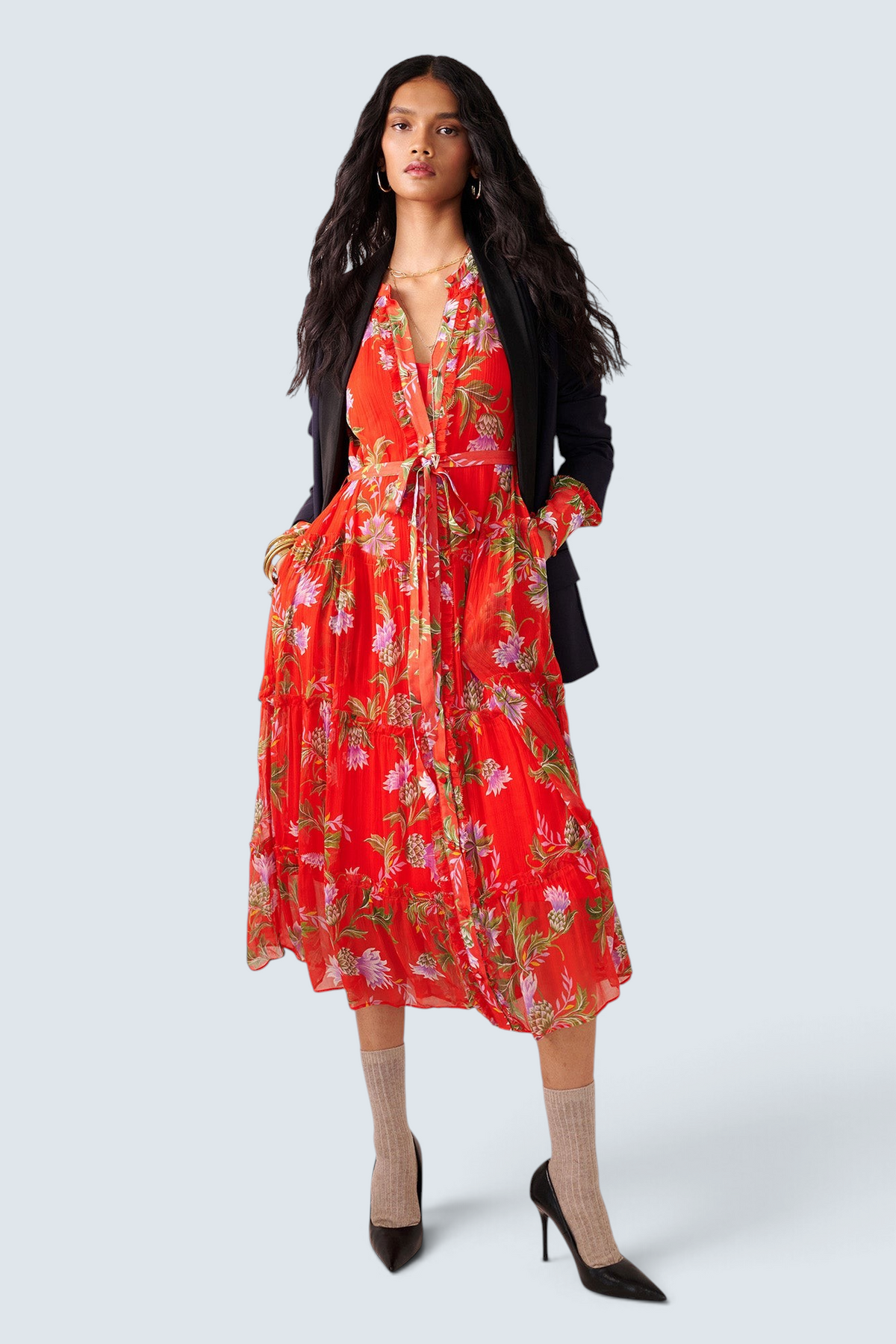 Front view of woman standing. She is wearing the Emira dress in the Stuart Thistle print. It is a flowing Flame-colored crinkle chiffon dress with sheer sleeves, delicate ruffles, fabric-covered buttons, and a detachable belt, featuring floral motifs inspired by the Scottish Highlands. Styled with a black blazer, socks and heels.