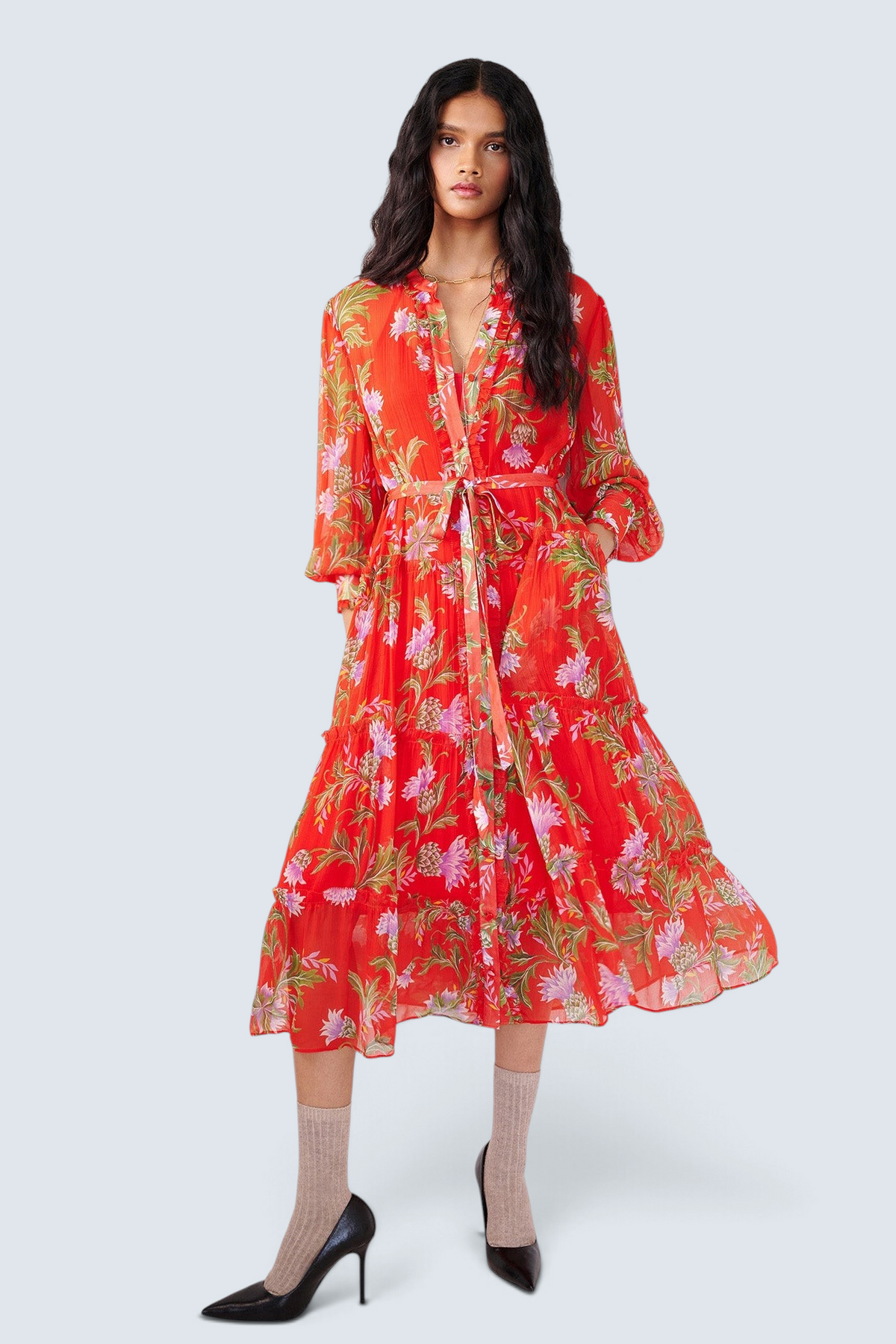 Front view of woman standing with a hand in pocket. She is wearing the Emira dress in the Stuart Thistle print. It is a flowing Flame-colored crinkle chiffon dress with sheer sleeves, delicate ruffles, fabric-covered buttons, and a detachable belt, featuring floral motifs inspired by the Scottish Highlands.