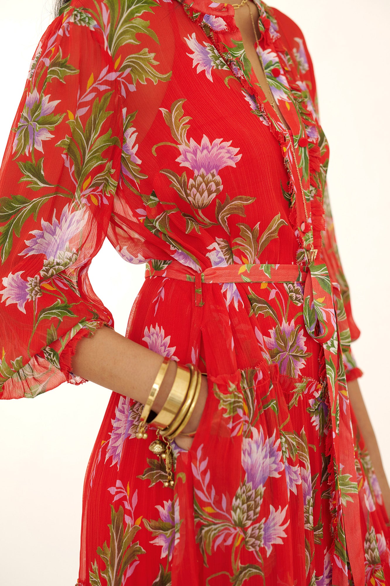 Side view close up of woman standing with hand in pocket. She is wearing the Emira dress in the Stuart Thistle print. It is a flowing Flame-colored crinkle chiffon dress with sheer sleeves, delicate ruffles, fabric-covered buttons, and a detachable belt, featuring floral motifs inspired by the Scottish Highlands. 