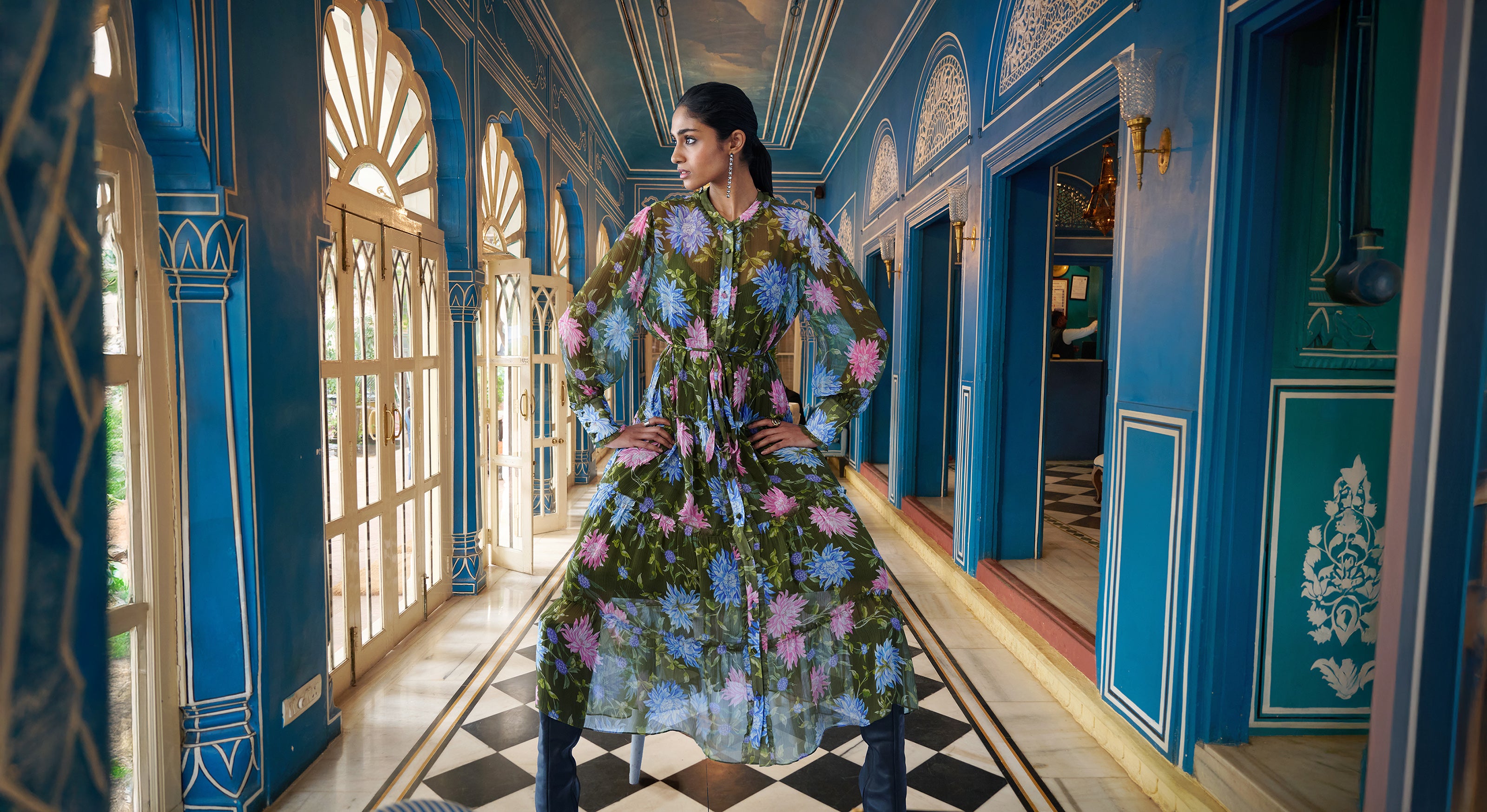 A woman is sitting in a conservatory. She is wearing an Emira Dress by Banjanan. The Emira Dress is crafted from crinkle chiffon. Its sheer sleeves, drapey skirt, and delicate ruffles create enchanting movement, with fabric-covered buttons, elevated eyelash details, and a detachable belt. It is in a Dahlia print with green background and is  finished with side-seam pockets. Slip included.
