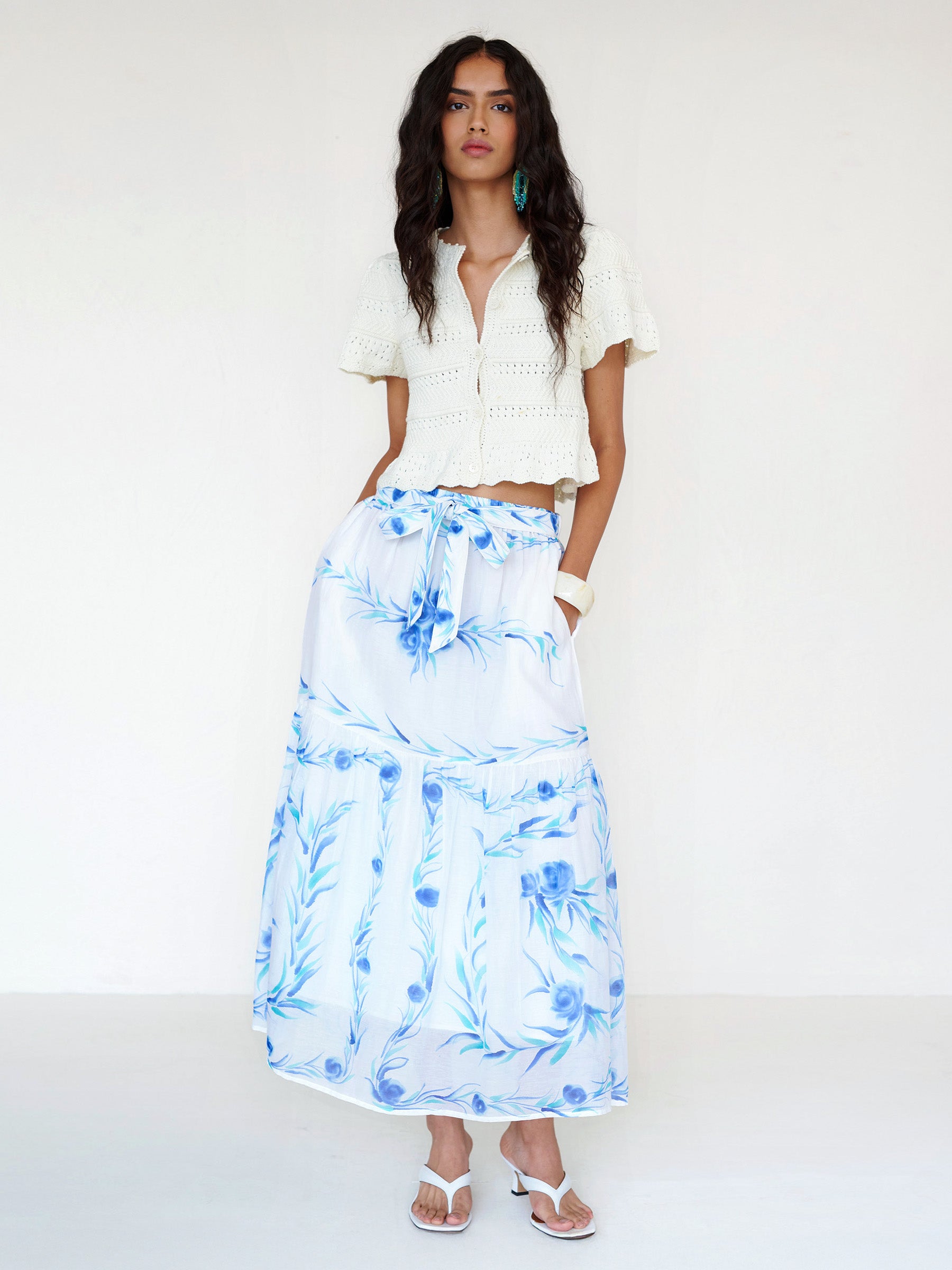 Front view of a woman standing. She is wearing the Flora full-length skirt with a hand-painted undulating floral print paired with a crochet cardigan. It features a tie at the waist and side-seam pockets.