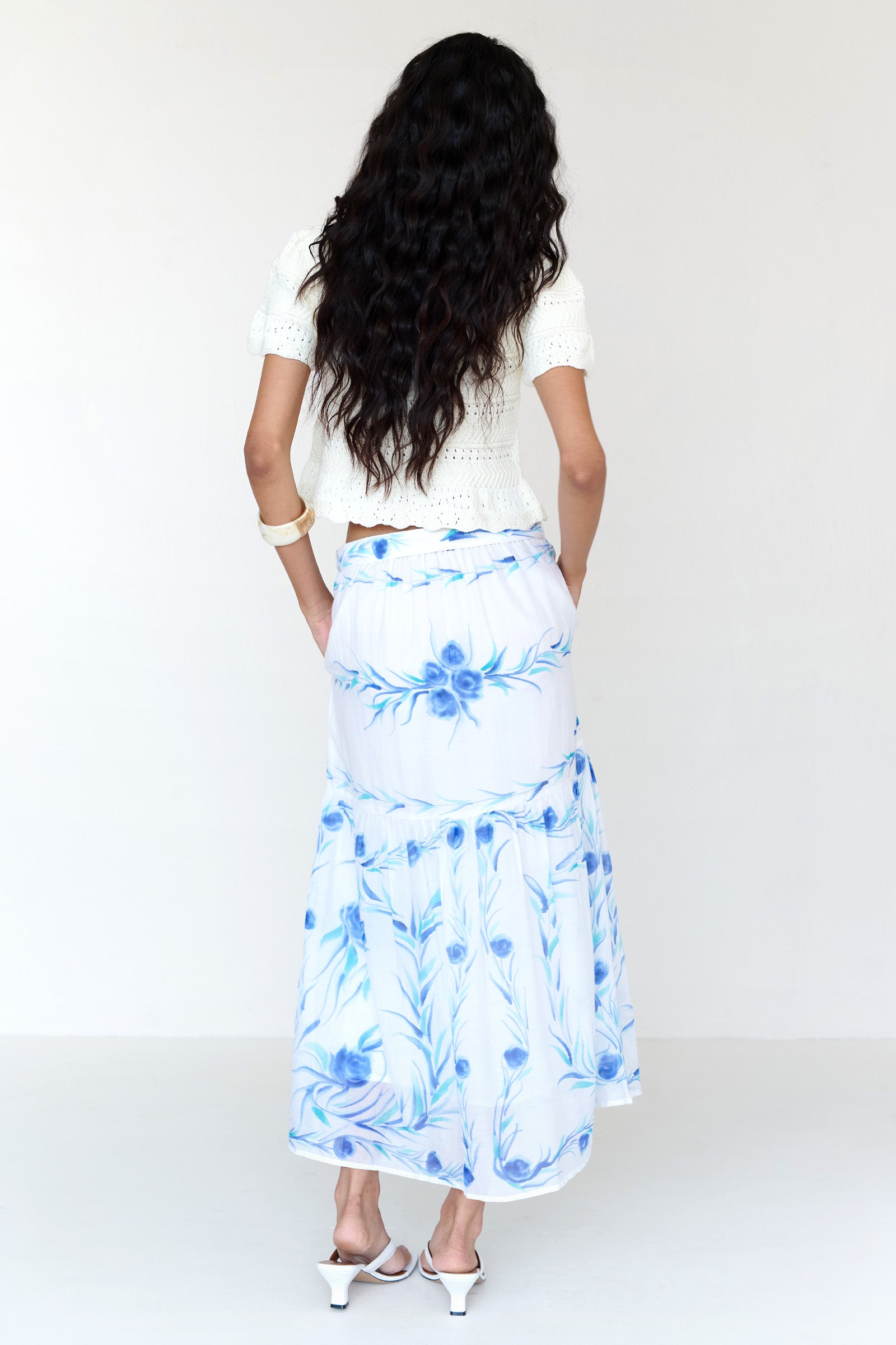 Back view of a woman standing. She is wearing the Flora skirt, featuring a hand-painted undulating floral print, full-length silhouette with waist tie, side-seam pockets, and full lining styled with a cardigan.