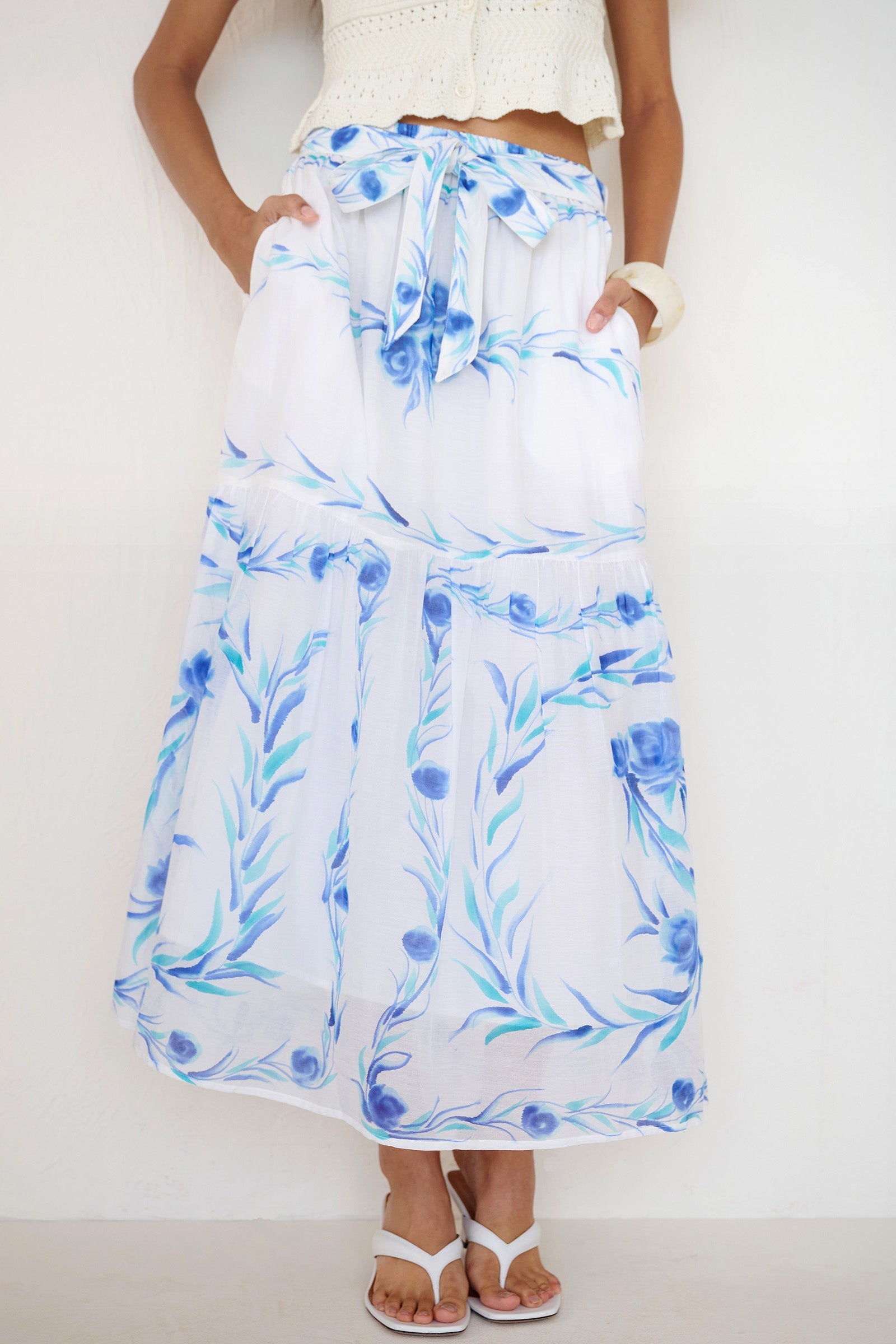 Front view close up of a woman standing with hands in pocket. She is wearing the Flora full-length skirt with a hand-painted undulating floral print, featuring a tie at the waist and side-seam pockets.