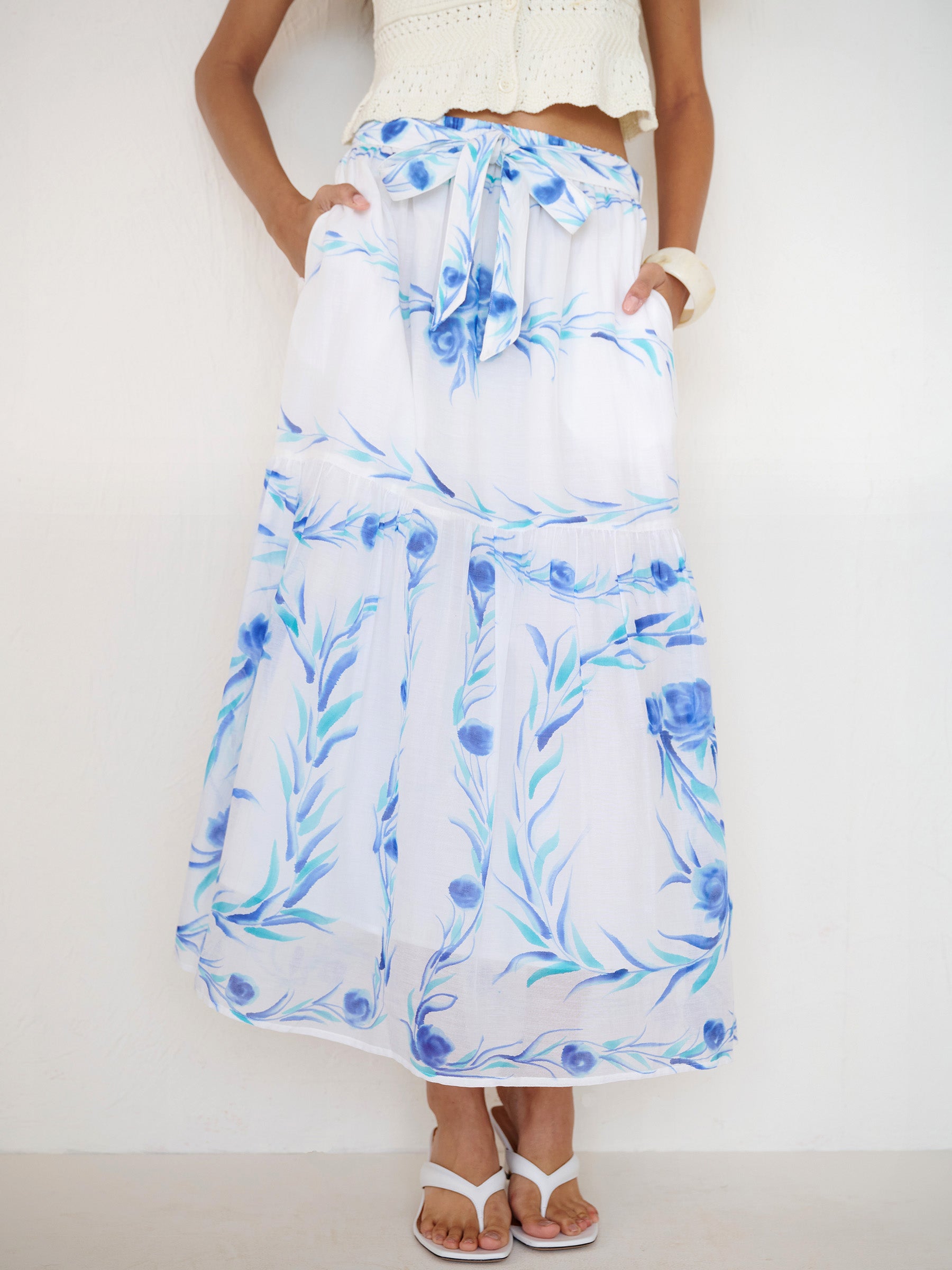 Front view close up of a woman standing with hands in pocket. She is wearing the Flora full-length skirt with a hand-painted undulating floral print, featuring a tie at the waist and side-seam pockets.