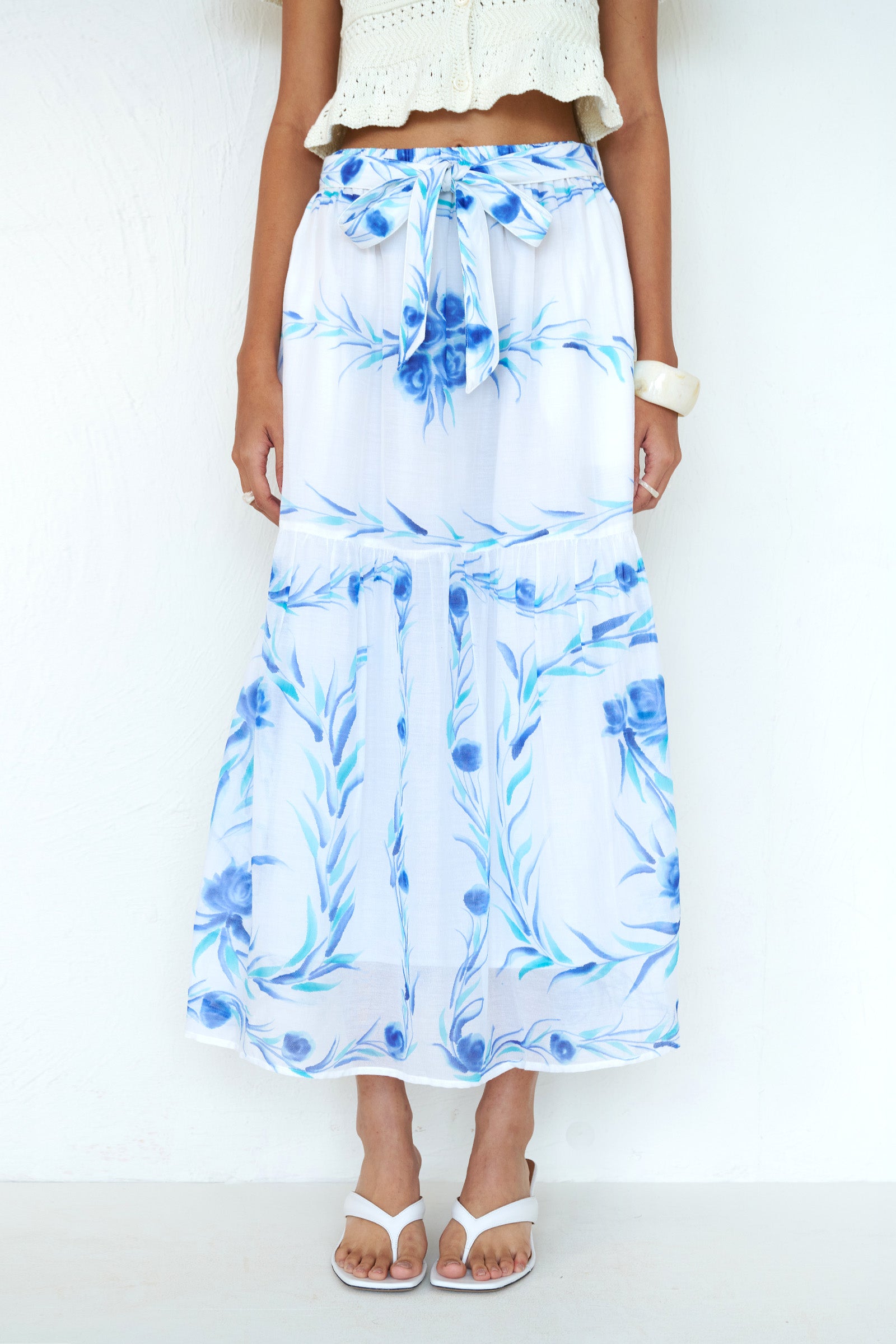 Front view close up of a woman standing. She is wearing the Flora skirt, featuring a hand-painted undulating floral print, full-length silhouette with waist tie, side-seam pockets, and full lining styled with a cardigan.