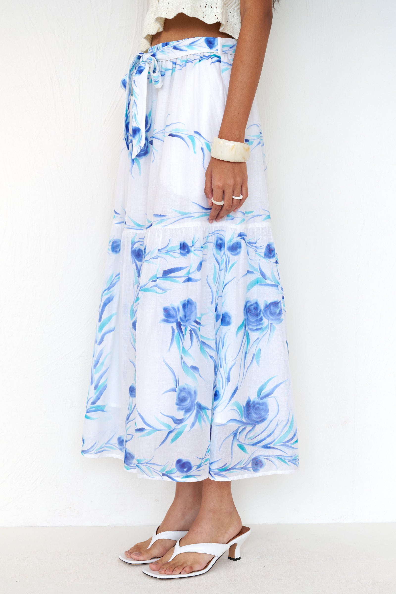 Side view close up of a woman standing. She is wearing the Flora skirt, featuring a hand-painted undulating floral print, full-length silhouette with waist tie, side-seam pockets, and full lining styled with a cardigan.