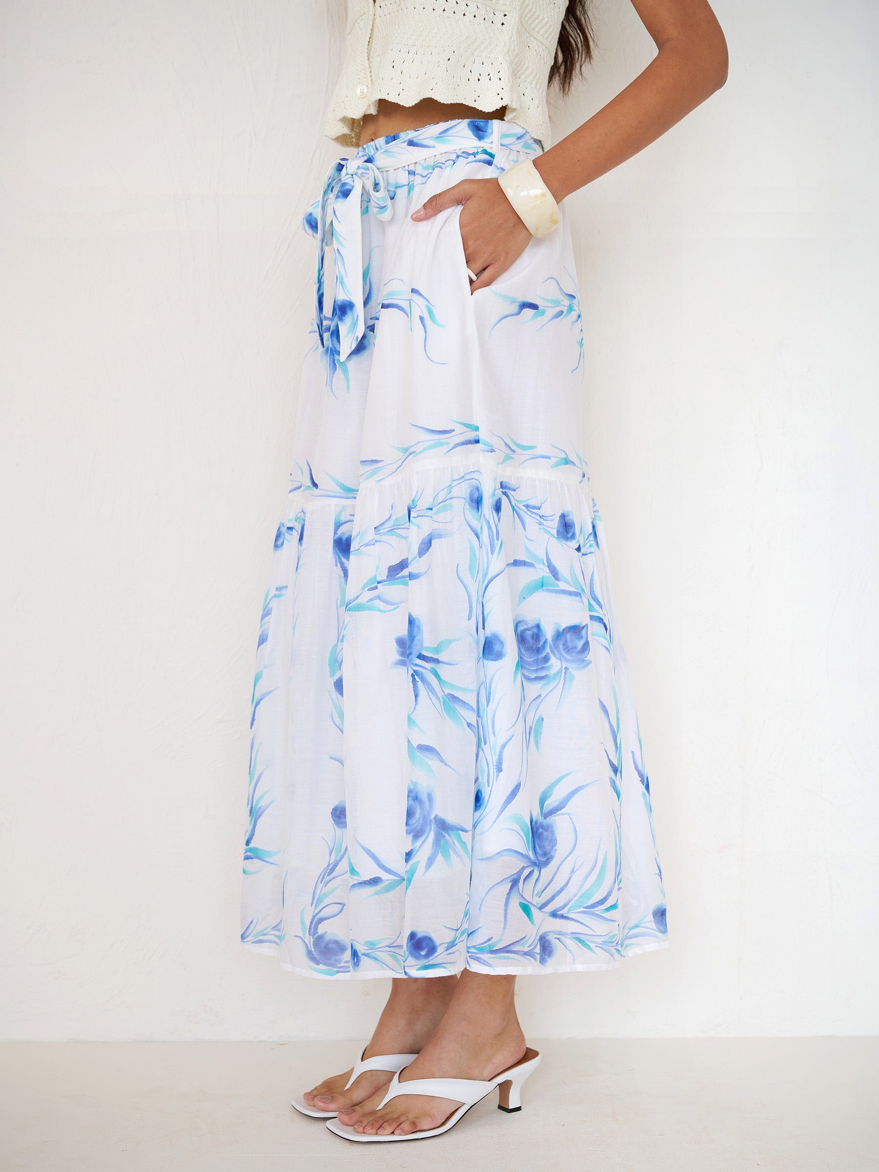 Side view close up of a woman standing with hands in pocket. She is wearing the Flora full-length skirt with a hand-painted undulating floral print, featuring a tie at the waist and side-seam pockets.