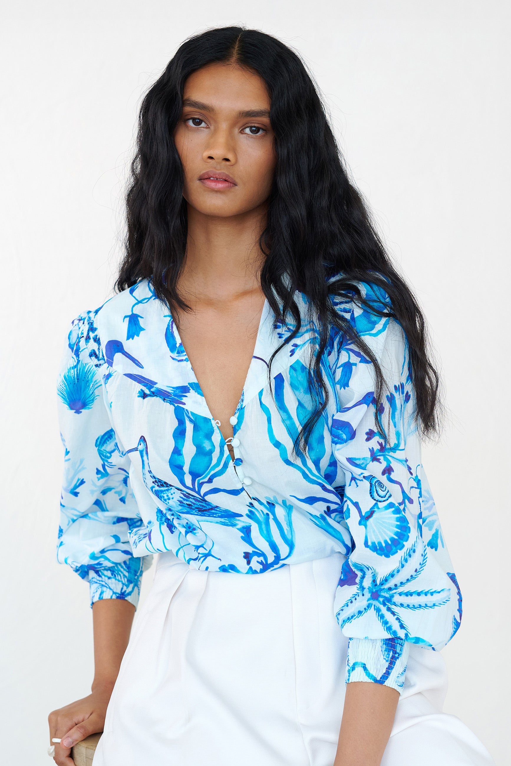 Front view of a woman sitting on a chair. She is wearing the Florrie shirt in Tidepool print. It features a V-neck yolk seam, fabric-covered buttons with elastic loops, smocked sleeves, and tonal underwater blue motifs.