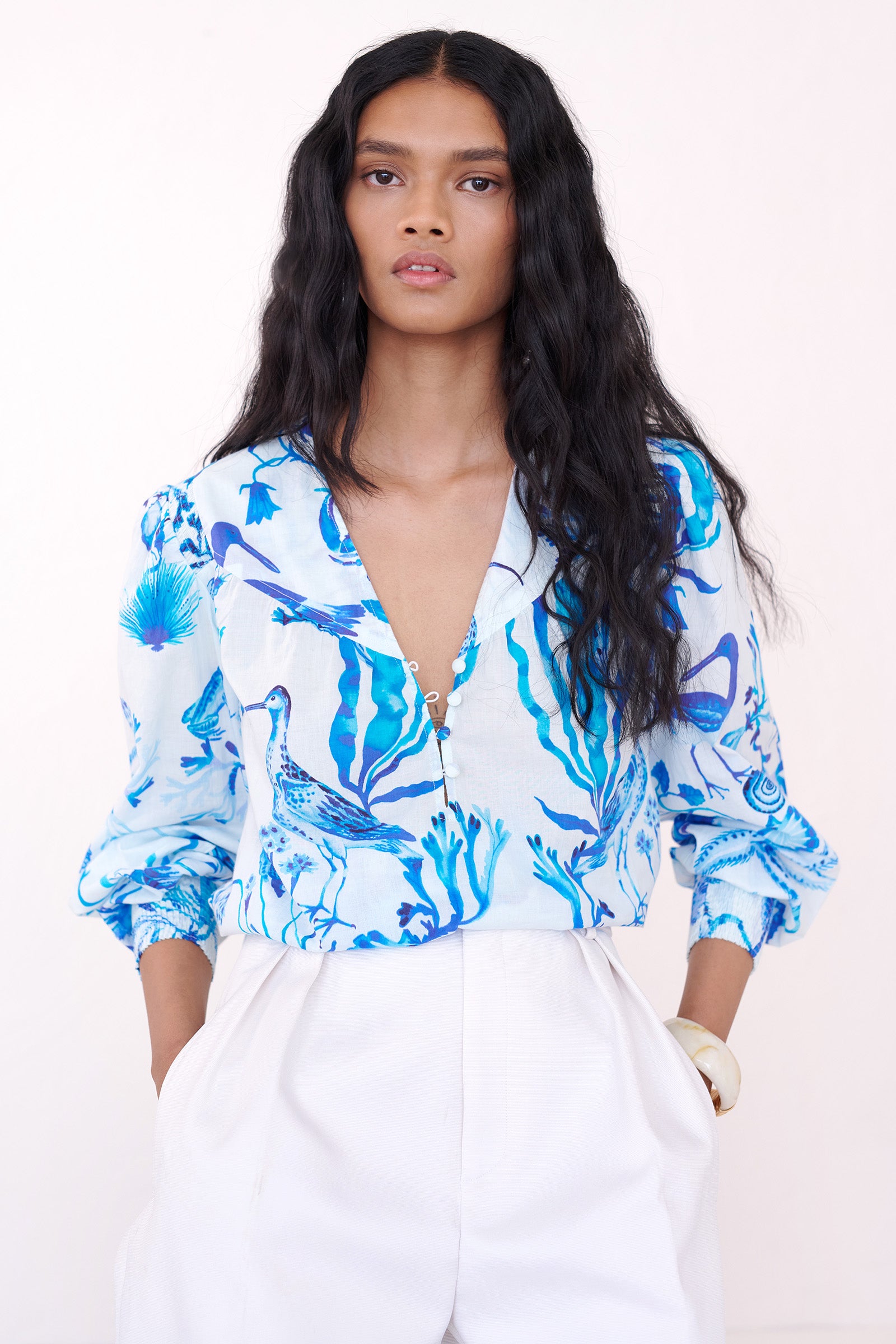 Front view of a woman ssitting on a chair. She is wearing the Florrie shirt in Tidepool print, featuring a V-neck yoke, fabric-covered buttons, and softly smocked sleeves in lightweight cotton voile, styled with white shorts.