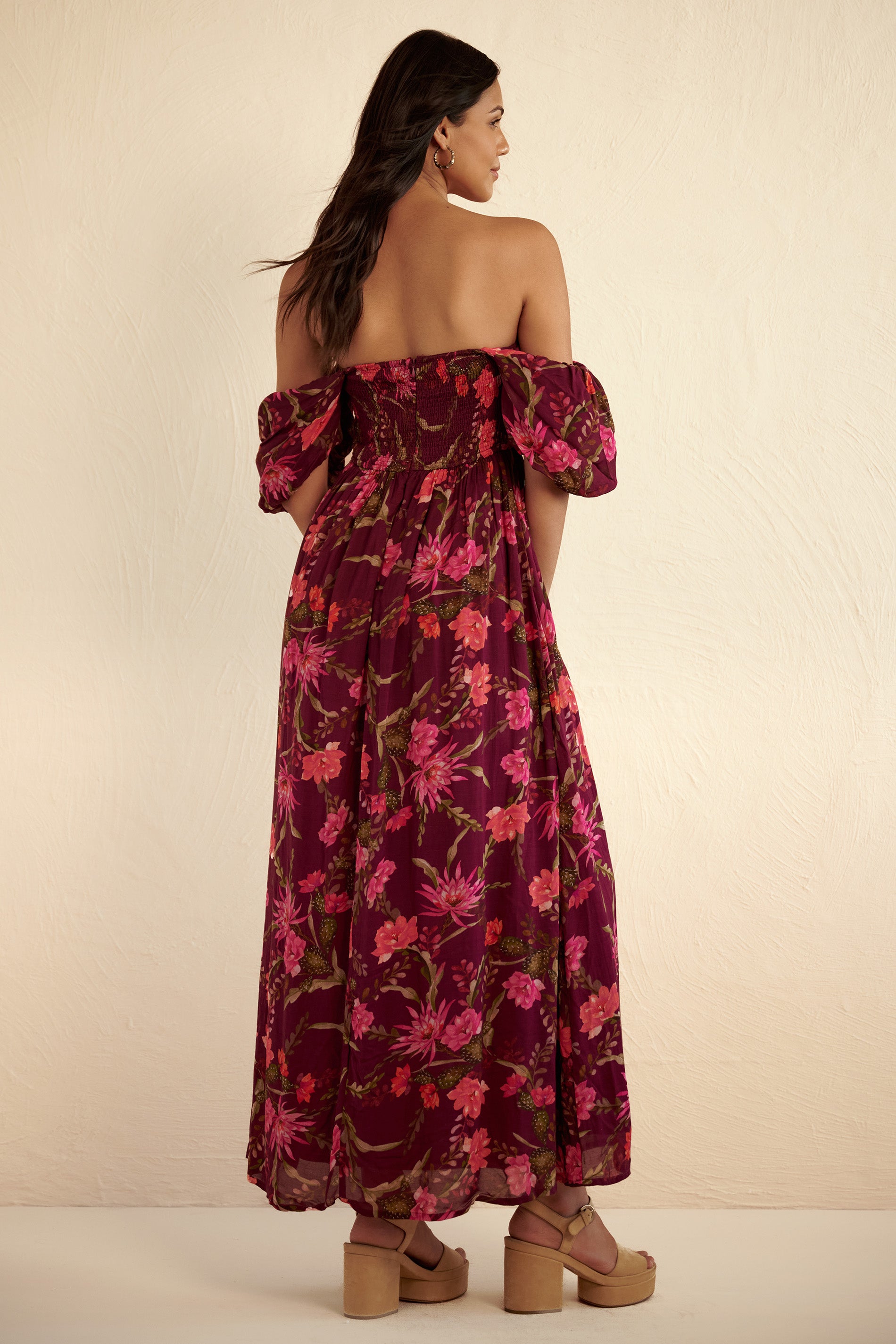 Back view of woman wearing the Farida dress in dusk magenta with watercolor-style cactus floral print, featuring a ruched bodice with boning, smocked back, off-shoulder elasticated sleeves, full-length skirt, and side pockets.