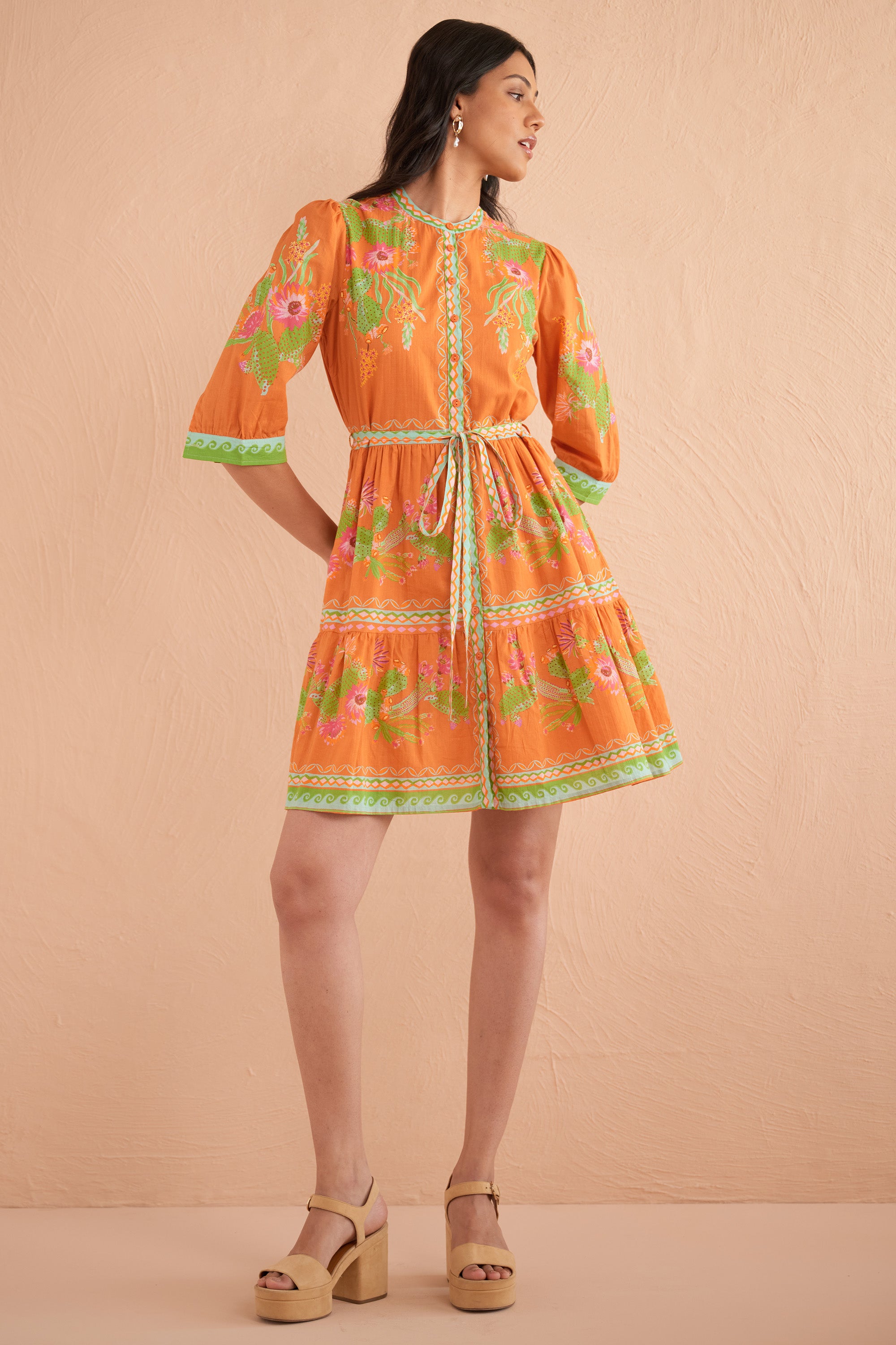 Front view of woman wearing Gizela Dress in Calypso Cacti Caramel print, a round-neck shirtdress with buttoned cuffs, self-tie waist belt, and side-seam pockets in sun-drenched cactus blossom cotton voile.