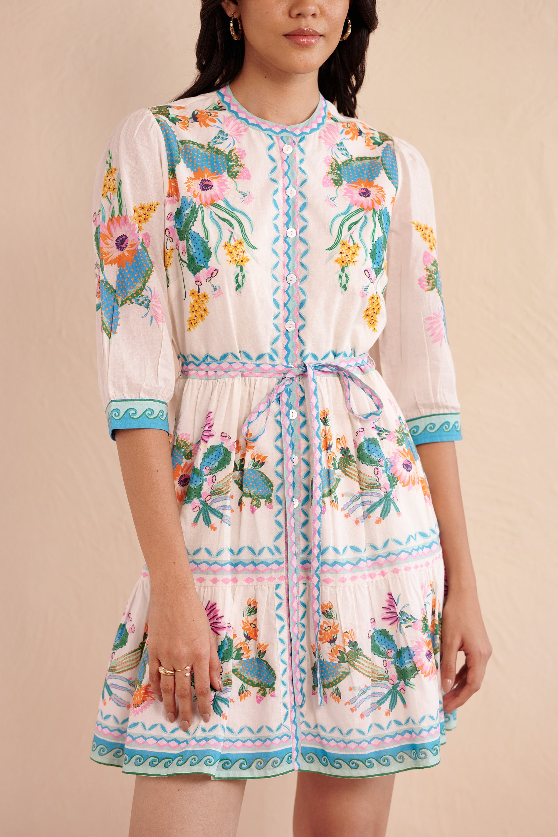 Close up of woman wearing a white knee-length dress with colorful Calypso Cacti Paradise print featuring blooming cacti and flowers, detailed with blue and pink geometric borders, 3/4 sleeves, and a drawstring waist.