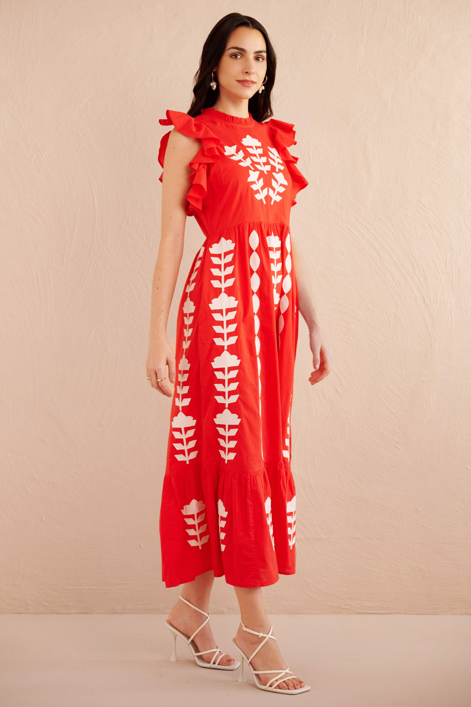 Front side view of woman wearing a sleeveless red midi dress with white appliqué floral embroidery, ruffled shoulders, and tiered hem, styled with white strappy heels.