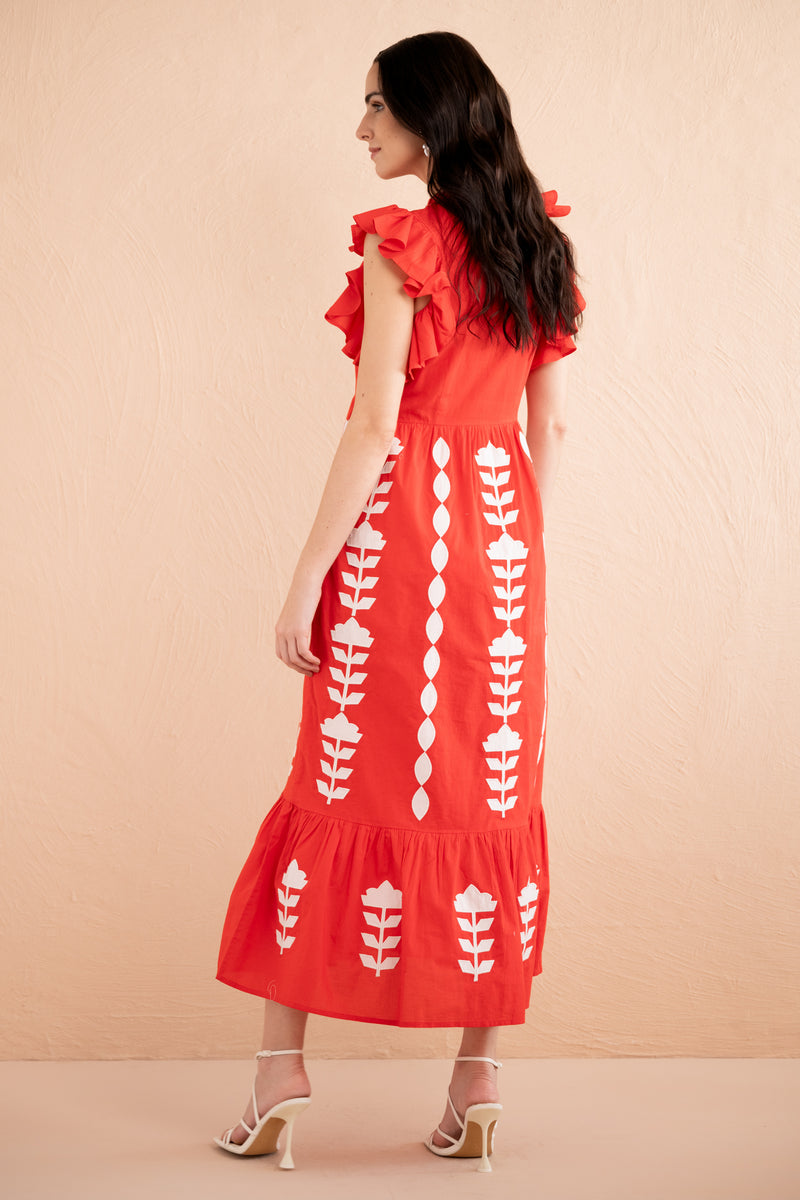 Back view of a woman wearing a sleeveless red midi dress with white appliqué embroidery in vertical floral patterns, ruffled shoulders, and a tiered hem.