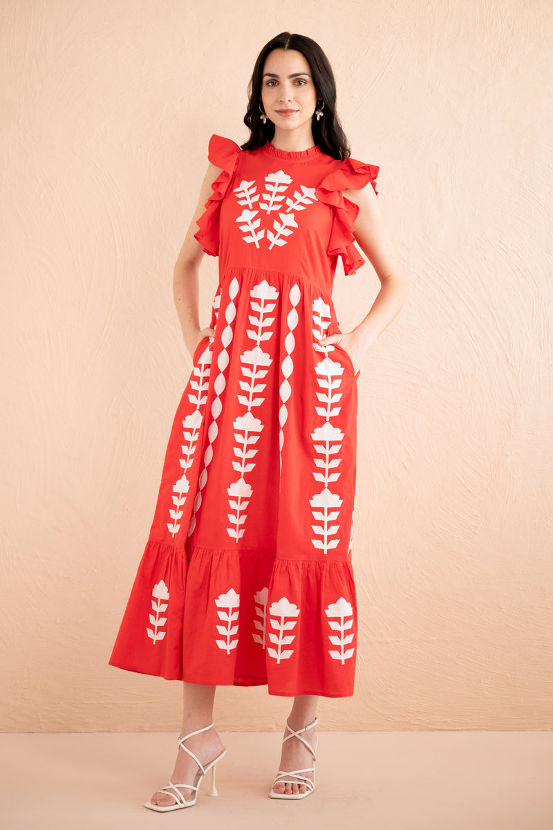 Front view of a woman wearing a sleeveless red midi dress with white appliqué embroidery in vertical floral patterns, ruffled shoulders, and a tiered hem.