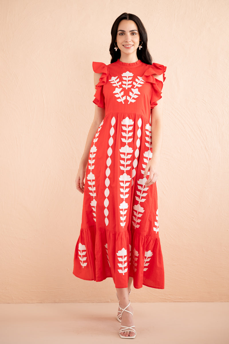 Front view of a woman wearing a sleeveless red midi dress with white appliqué embroidery in vertical floral patterns, ruffled shoulders, and a tiered hem.