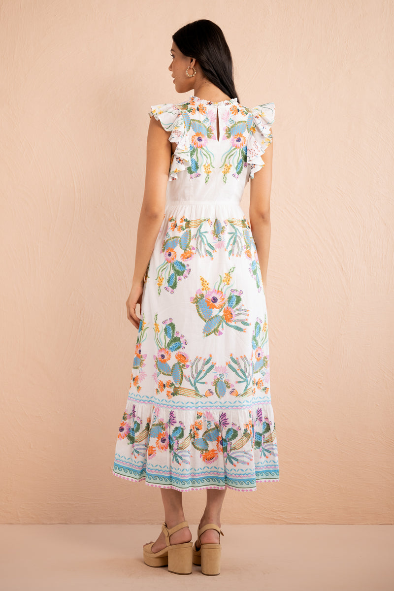 Back view of a woman wearing a sleeveless white midi dress with colorful Calypso Cacti Paradise print, ruffled shoulders, a fitted waist, and a flared tiered hem.