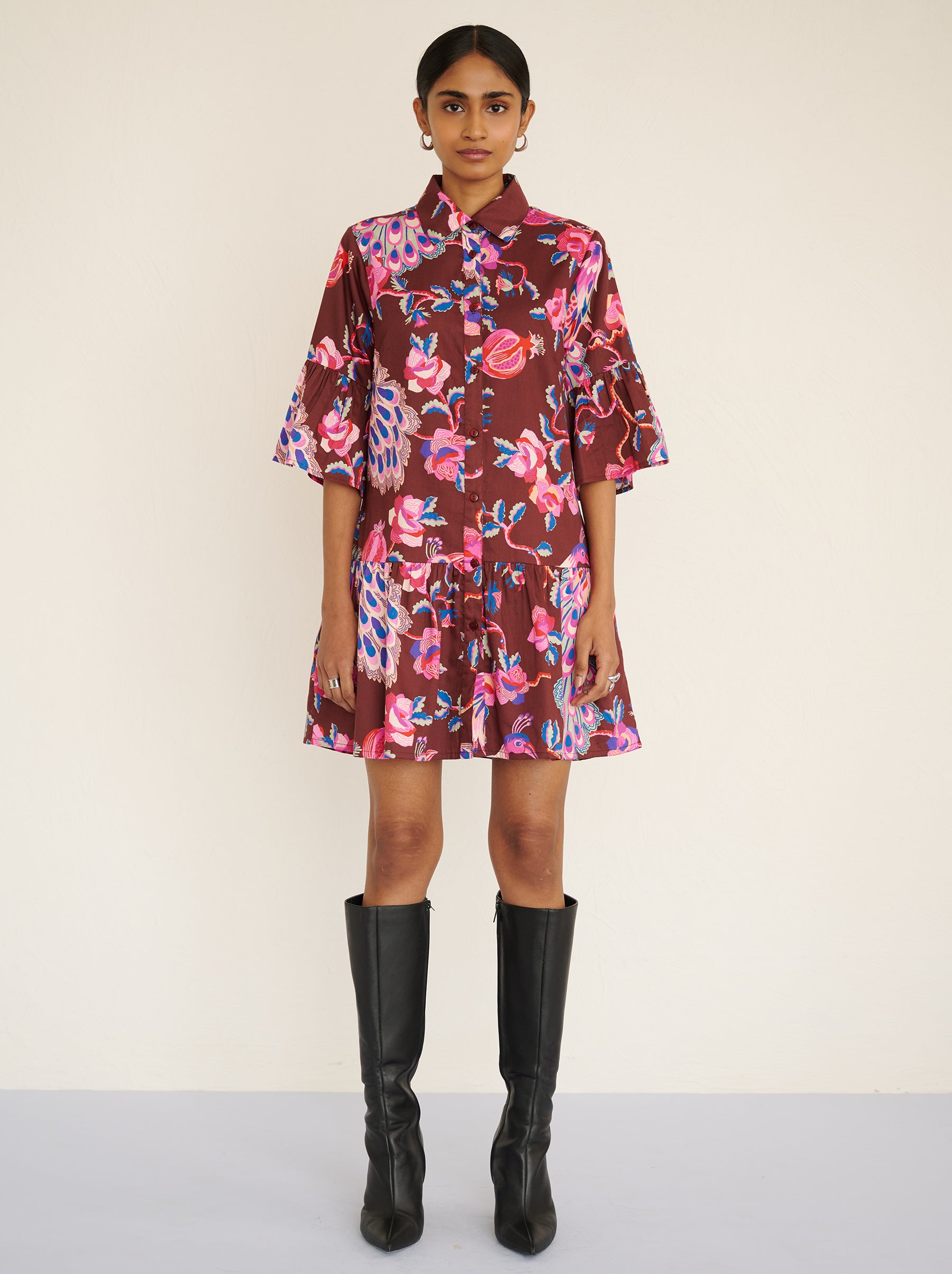 Front view of a model standing. She is wearing the Hydra dress in the Bukhara Peacock print in rum color. It is featuring ruffled sleeves and hem, fabric-covered buttons, and side-seam pockets.