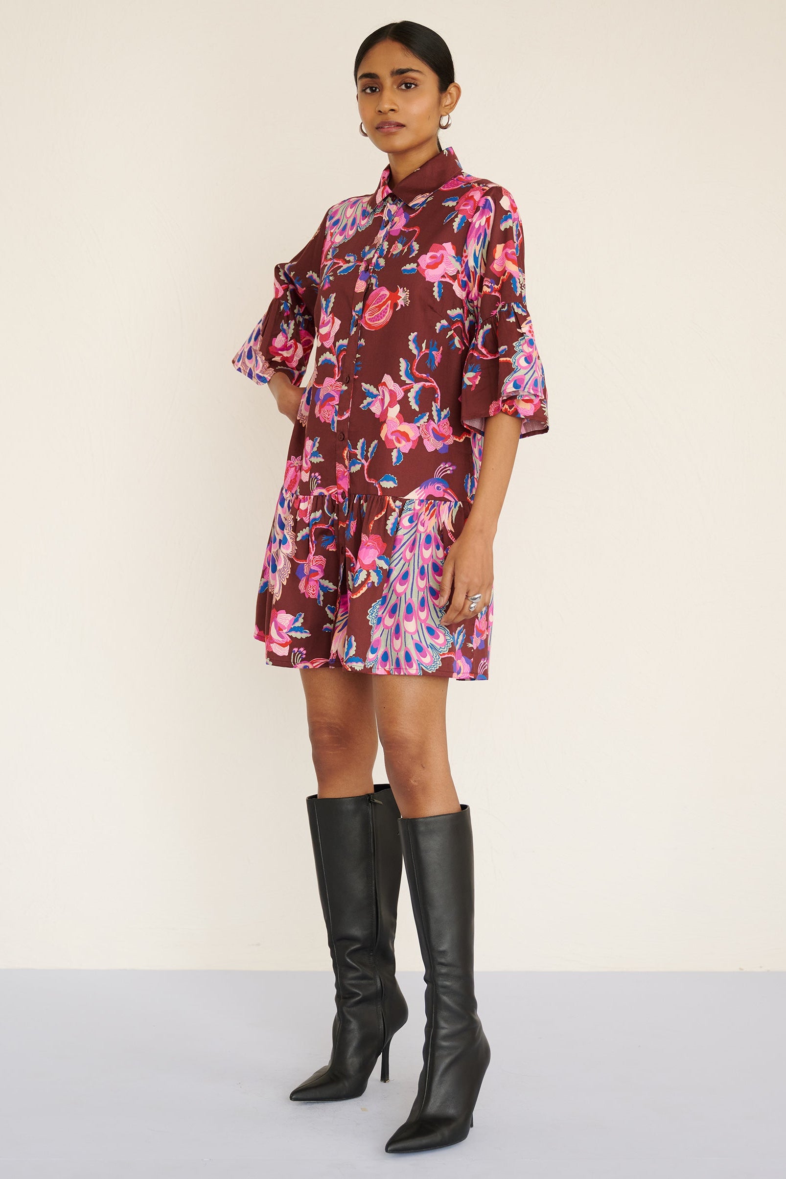 Side view of a model standing. She is wearing the Hydra dress in the Bukhara Peacock print in rum color. It is featuring ruffled sleeves and hem, fabric-covered buttons, and side-seam pockets.