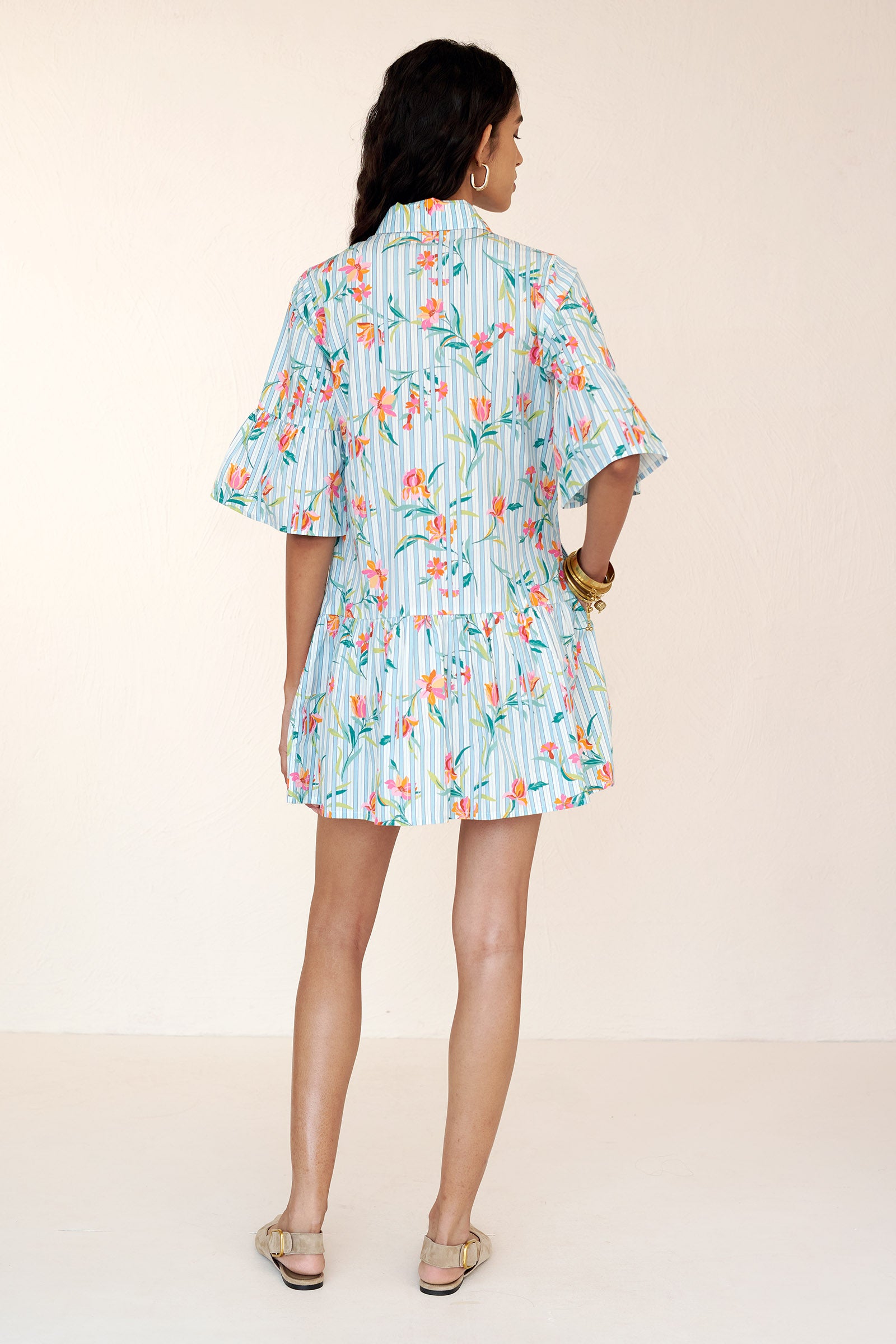 Back view of a woman standing wearing the Hydra mini dress in orchid stripe print. It features a button placket, slightly dropped waist, bell sleeves, side-seam pockets, and crisp cotton poplin.