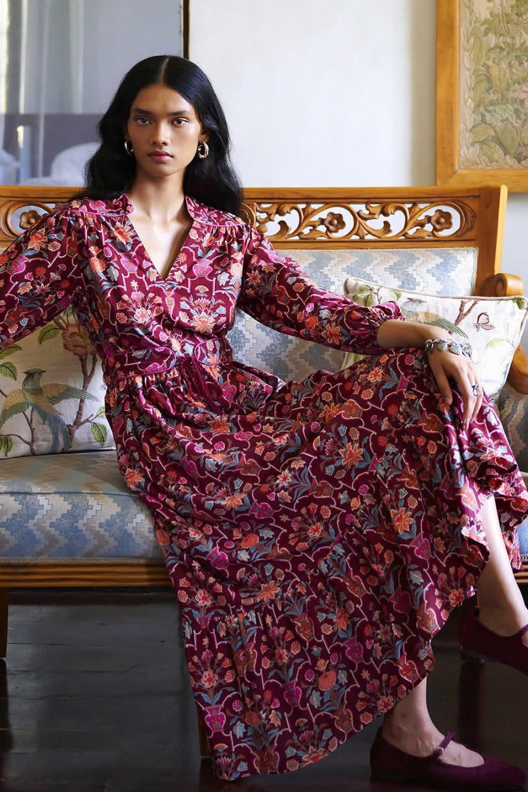 A woman wearing a long-sleeve wrap-style midi dress with a dense floral and abstract print in deep pink, burgundy, and muted blue, seated on a carved wooden bench with patterned cushions.