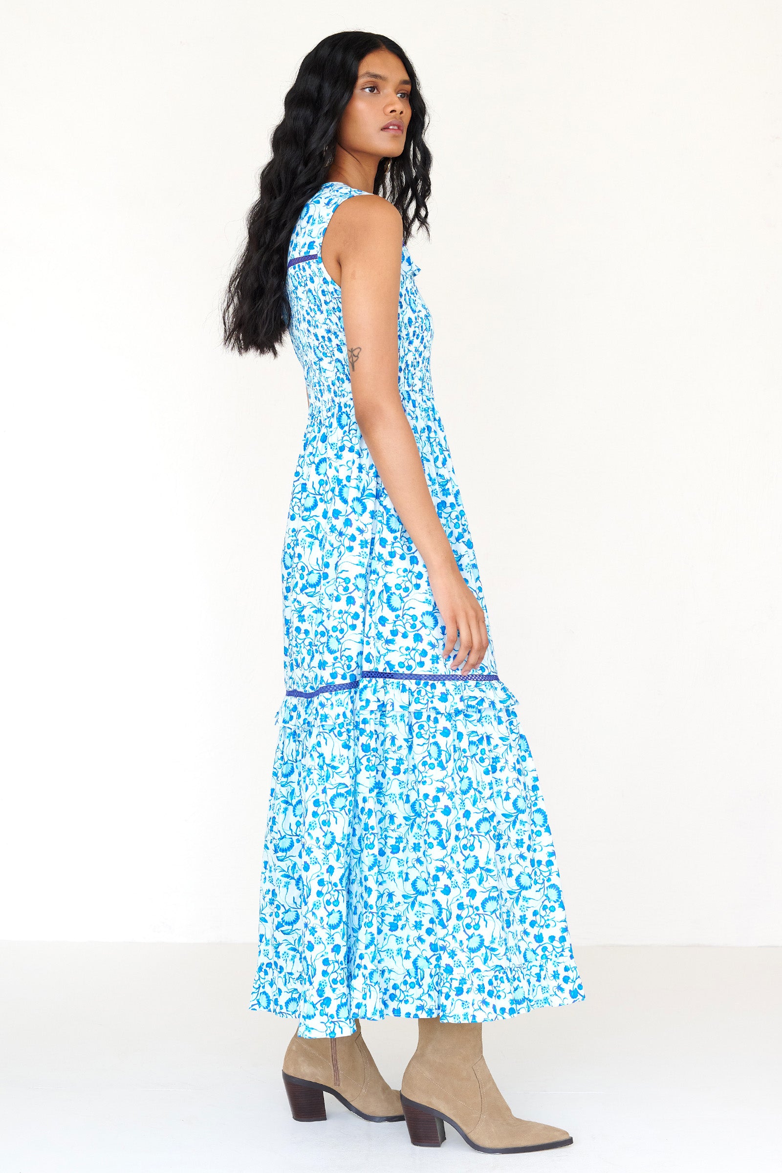 Side view of a woman standing wearing the Iris dress in Clam Garland print. It features a smocked bodice, tonal lace detailing, keyhole back with loop button and pockets.
