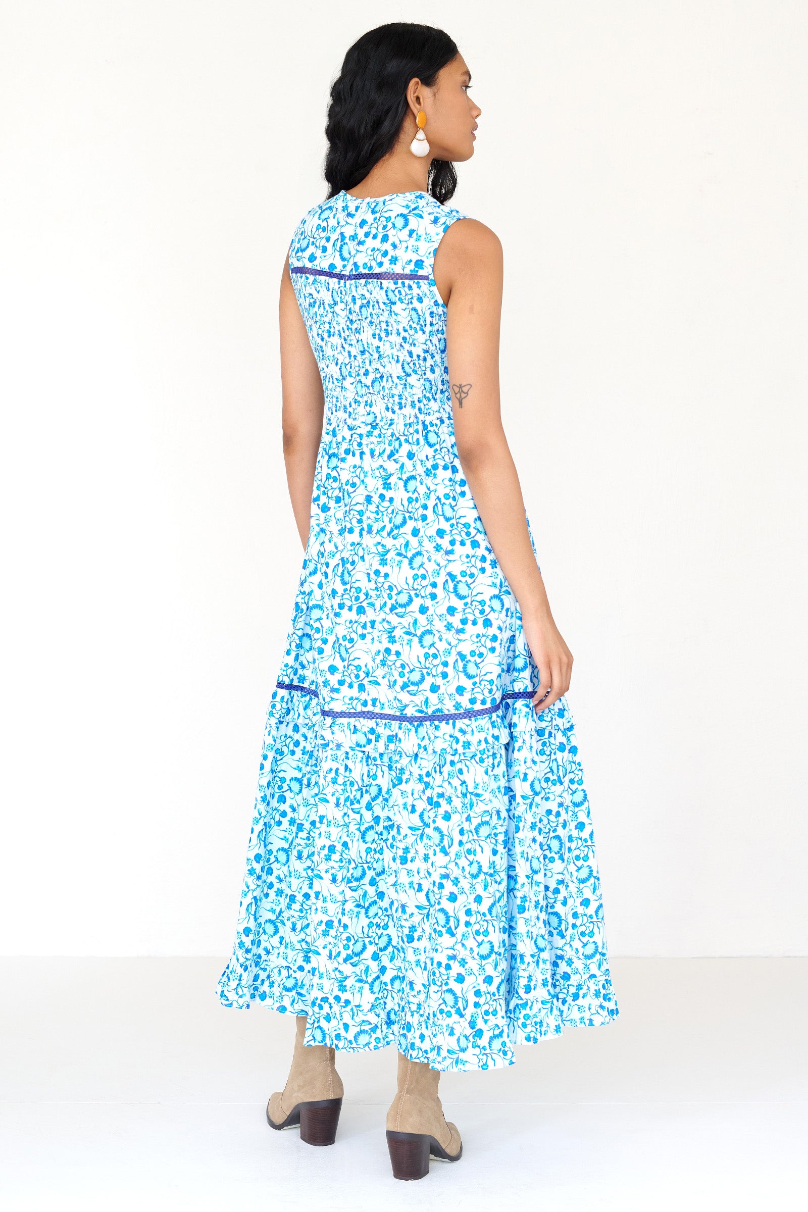 Back view of a woman standing wearing the Iris dress in Clam Garland print. It features a smocked bodice, tonal lace detailing, keyhole back with loop button and pockets.