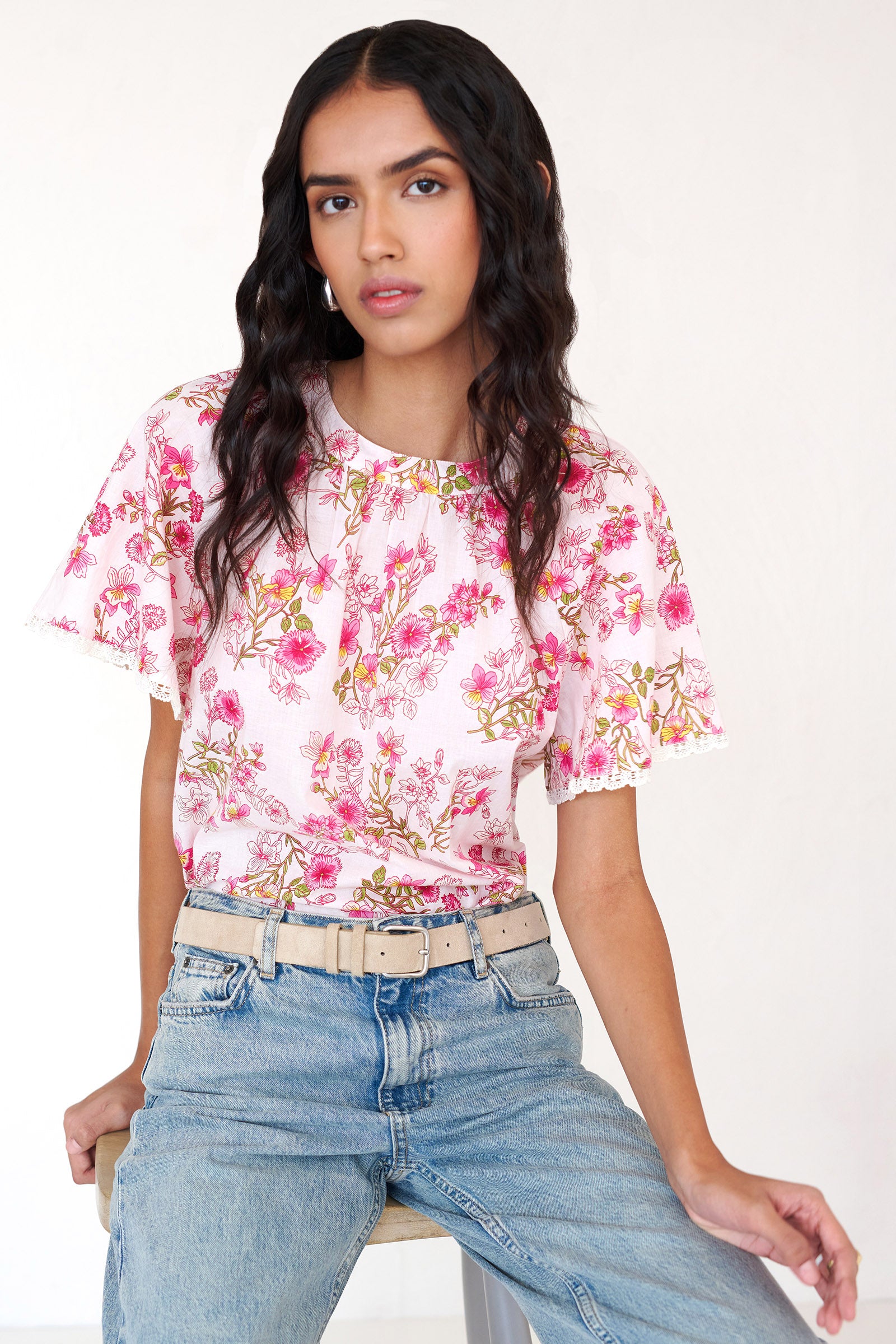 Front view of woman sitting on a chair wearing the Joyful top with tucked raglan sleeves, lace trim at the sleeves, and a Burdock wildflower print, finished with a buttoned keyhole at the back neck.