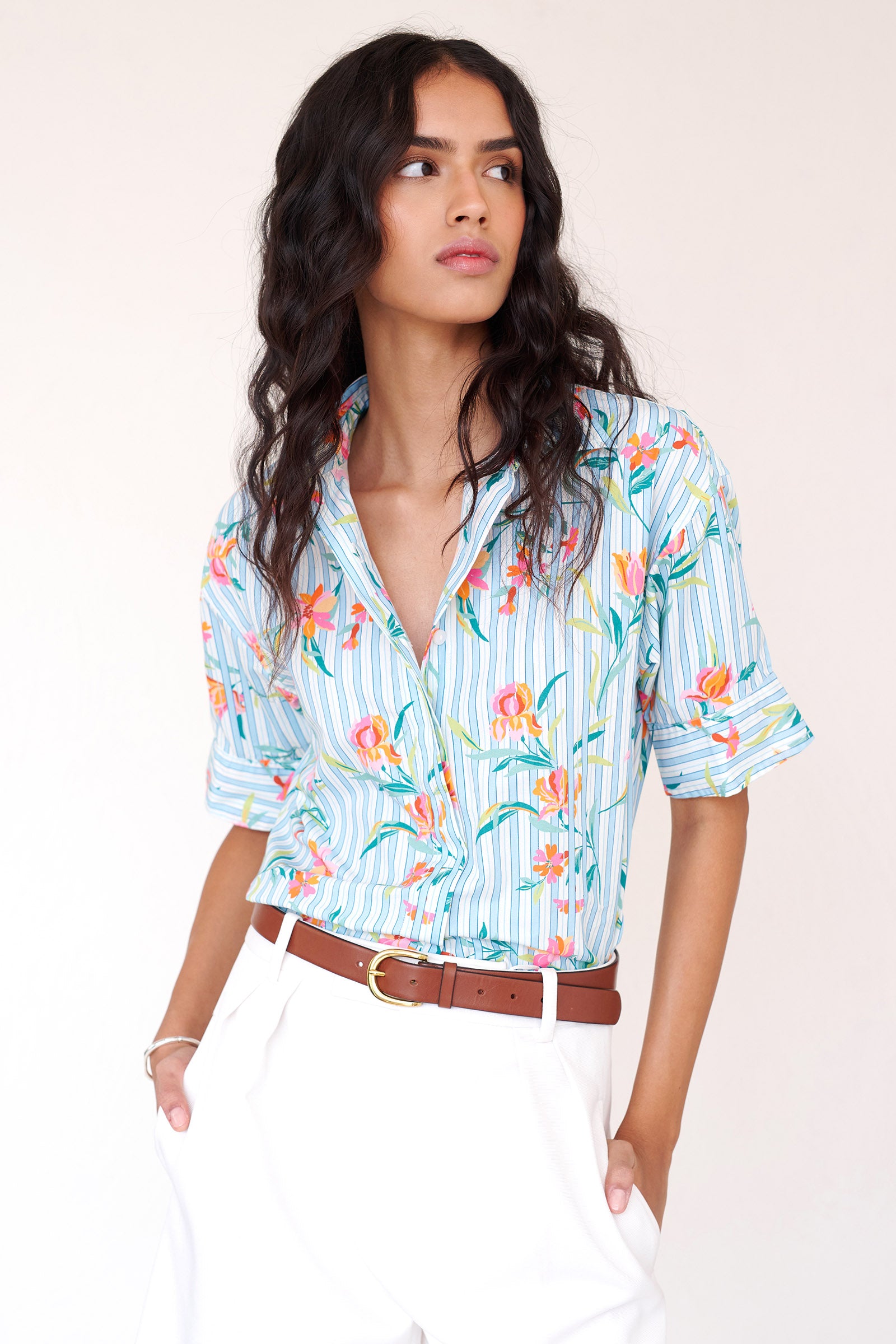 Front view close up of woman wearing the Jura shirt styled with white pants and brown belt. It features a fitted collared shirt with vented cuffs, mixed-direction pinstripes, and delicate orchid motifs.