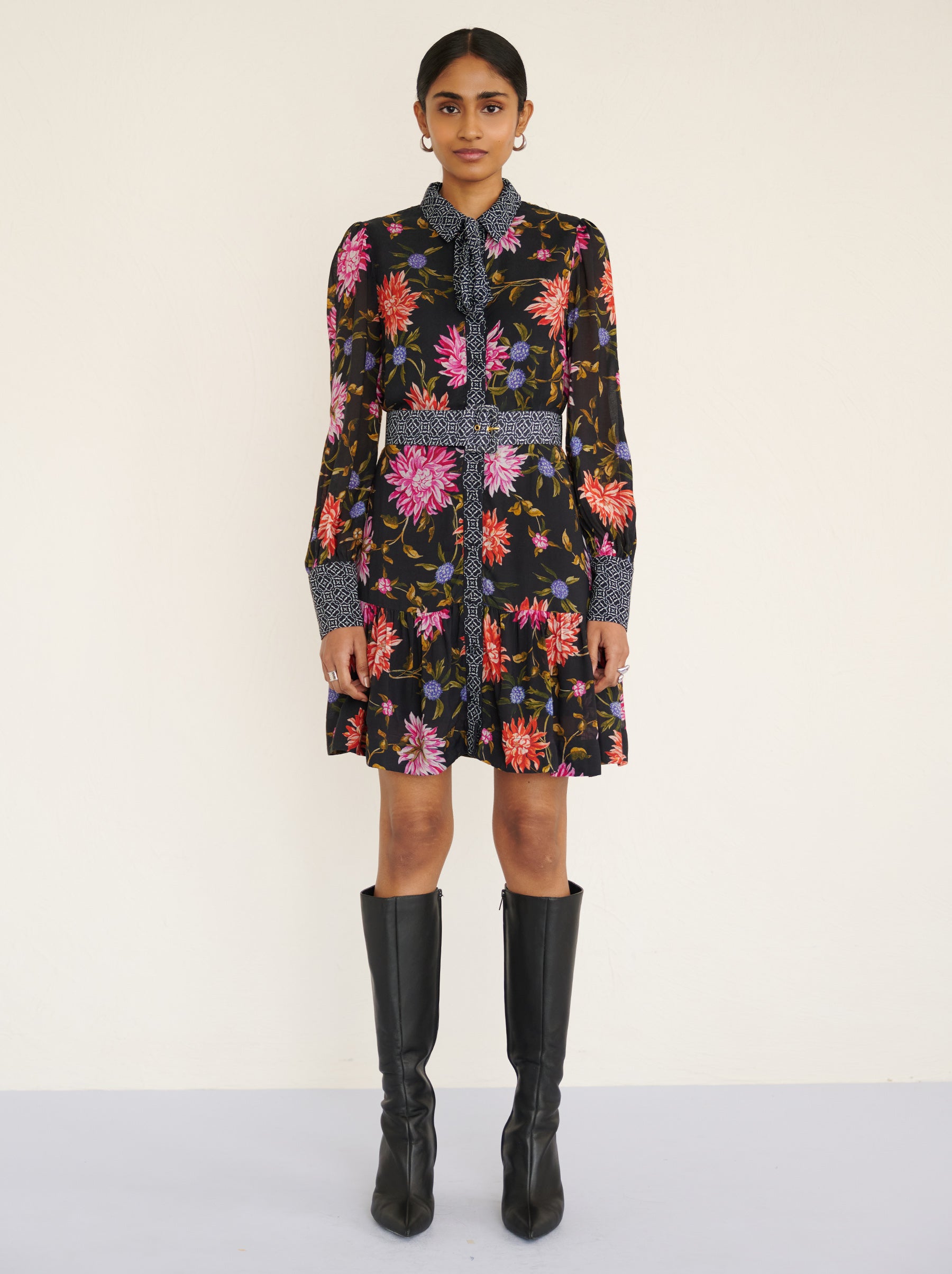 Front view of a model standing. She is wearing the Kay mini dress in lightweight viscose voile with the Dahlia floral print in Jet Black color with multicolor Dahlia. It is  featuring fabric-covered buttons, a bow at the neck, a fabric-covered belt, and side-seam pockets.