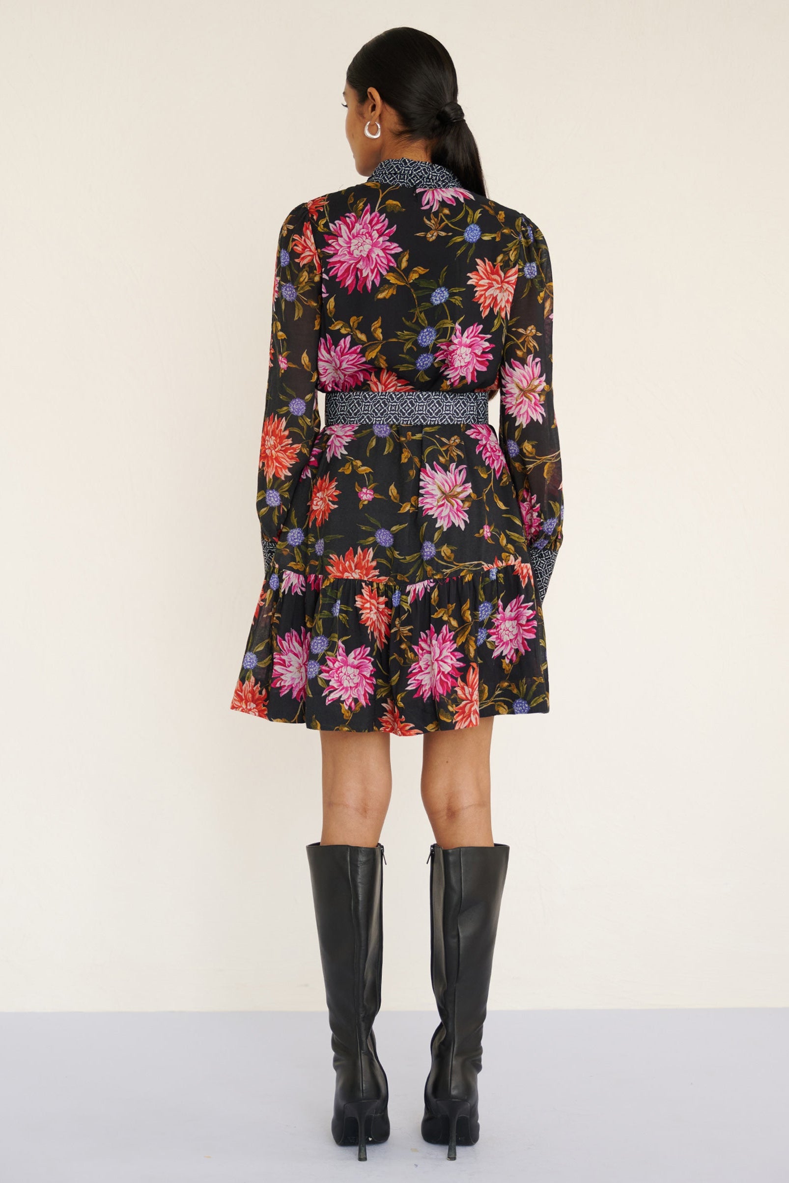 Back view of a model standing. She is wearing the Kay mini dress in lightweight viscose voile with the Dahlia floral print in Jet Black color with multicolor Dahlia. It is  featuring fabric-covered buttons, a bow at the neck, a fabric-covered belt, and side-seam pockets.