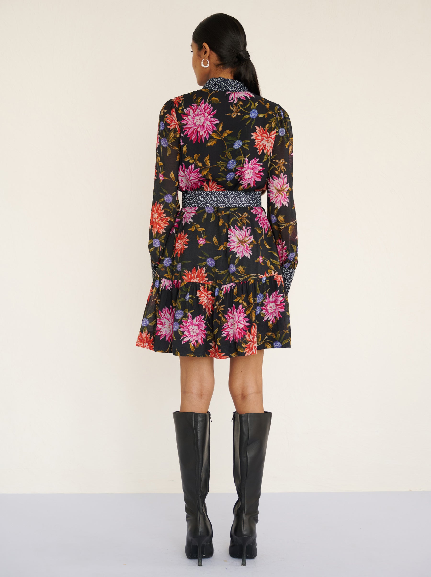 Back view of a model standing. She is wearing the Kay mini dress in lightweight viscose voile with the Dahlia floral print in Jet Black color with multicolor Dahlia. It is  featuring fabric-covered buttons, a bow at the neck, a fabric-covered belt, and side-seam pockets.