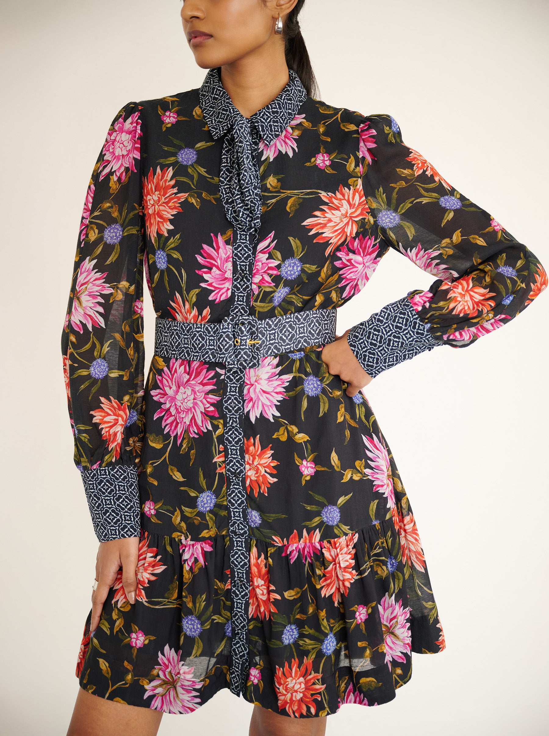 Front view close up of a model standing. She is wearing the Kay mini dress in lightweight viscose voile with the Dahlia floral print in Jet Black color with multicolor Dahlia. It is  featuring fabric-covered buttons, a bow at the neck, a fabric-covered belt, and side-seam pockets.