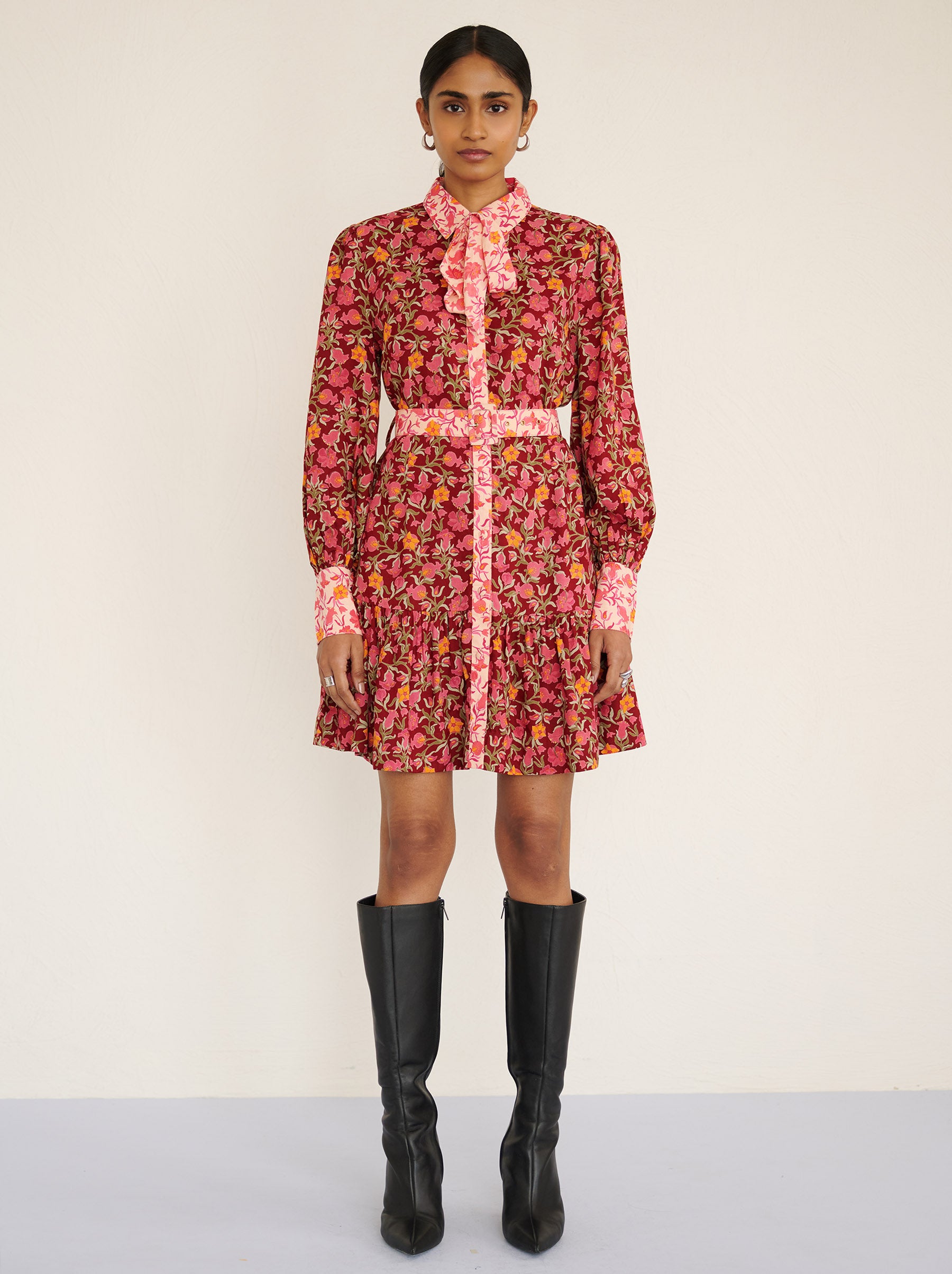 Front view of a model standing. She is wearing the Kay mini dress in eco-friendly viscose with the Pomegranate Vine print in Azalea color. It is featuring fabric-covered buttons, a bow at the neck, a fabric-covered belt, and side-seam pockets.
