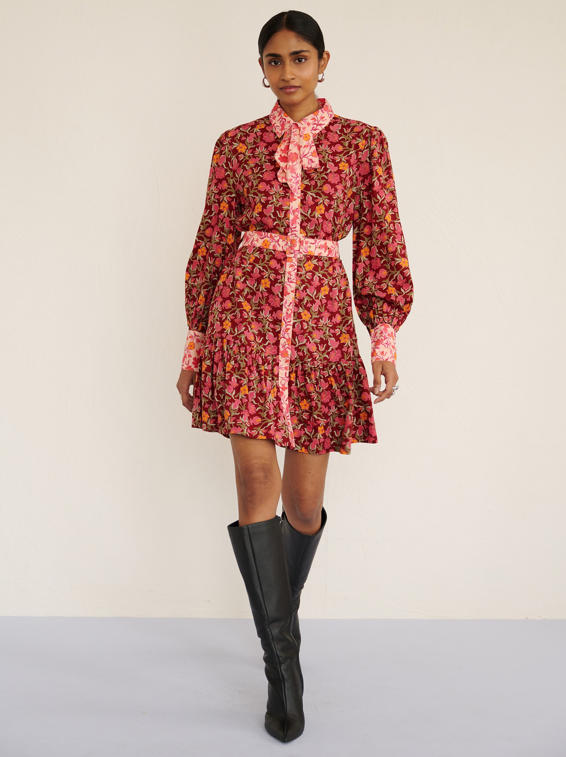Front view of a model walking. She is wearing the Kay mini dress in eco-friendly viscose with the Pomegranate Vine print in Azalea color. It is featuring fabric-covered buttons, a bow at the neck, a fabric-covered belt, and side-seam pockets.