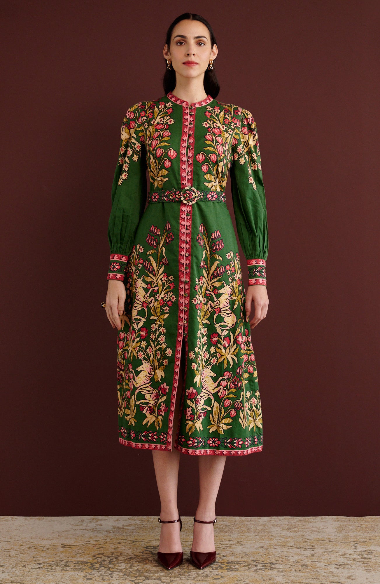 Front view of woman standing. She is wearing the Kay midi dress in Unicorn Tapestry print. It has a fitted bodice, stand collar, fabric-covered buttons, detachable belt, side-seam pockets, and 15th-century-inspired enchanted garden motifs on the shoulders and skirt.