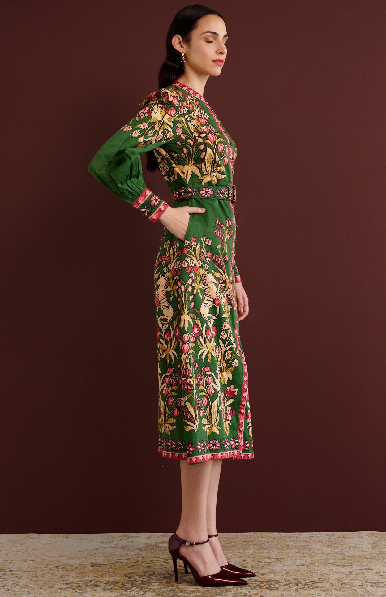 Side view of woman standing with her right hand in pocket. She is wearing the Kay midi dress in Unicorn Tapestry print. It has a fitted bodice, stand collar, fabric-covered buttons, detachable belt, side-seam pockets, and 15th-century-inspired enchanted garden motifs on the shoulders and skirt.