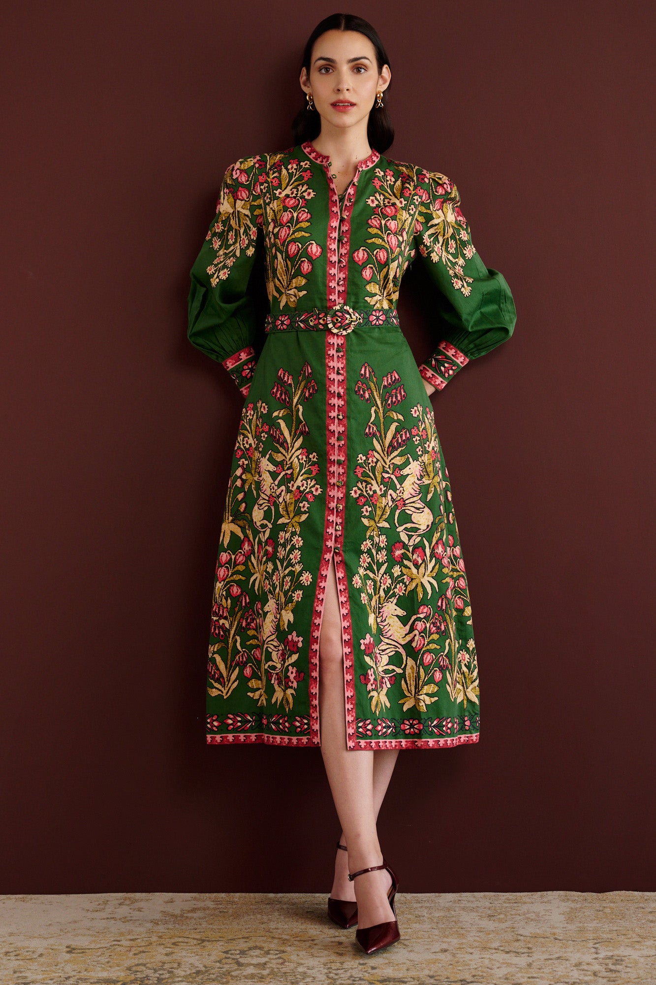 Front view of woman standing. She is wearing the Kay midi dress in Unicorn Tapestry print. It has a fitted bodice, stand collar, fabric-covered buttons, detachable belt, side-seam pockets, and 15th-century-inspired enchanted garden motifs on the shoulders and skirt.