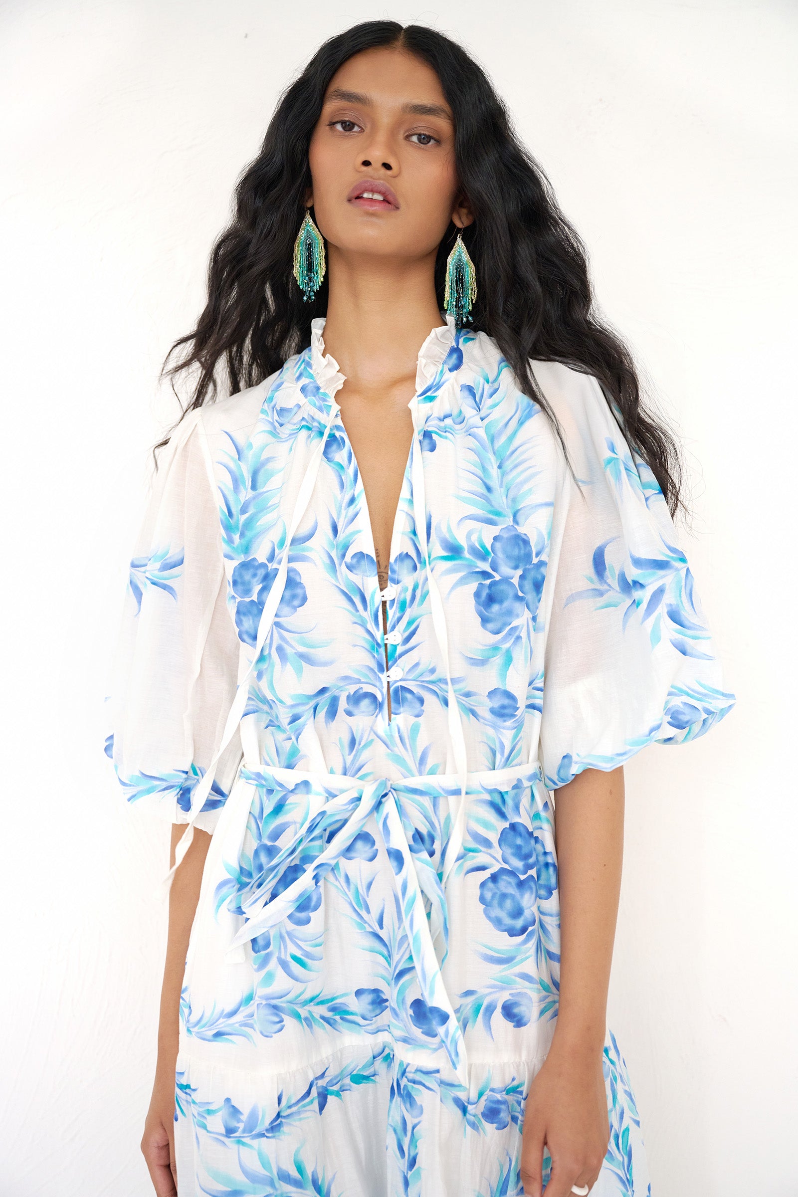 Front view close up of a woman standing wearing the Liza dress in cotton silk. It features hand-painted floral print, airy silhouette with fabric belt, tie and ruffled neckline.