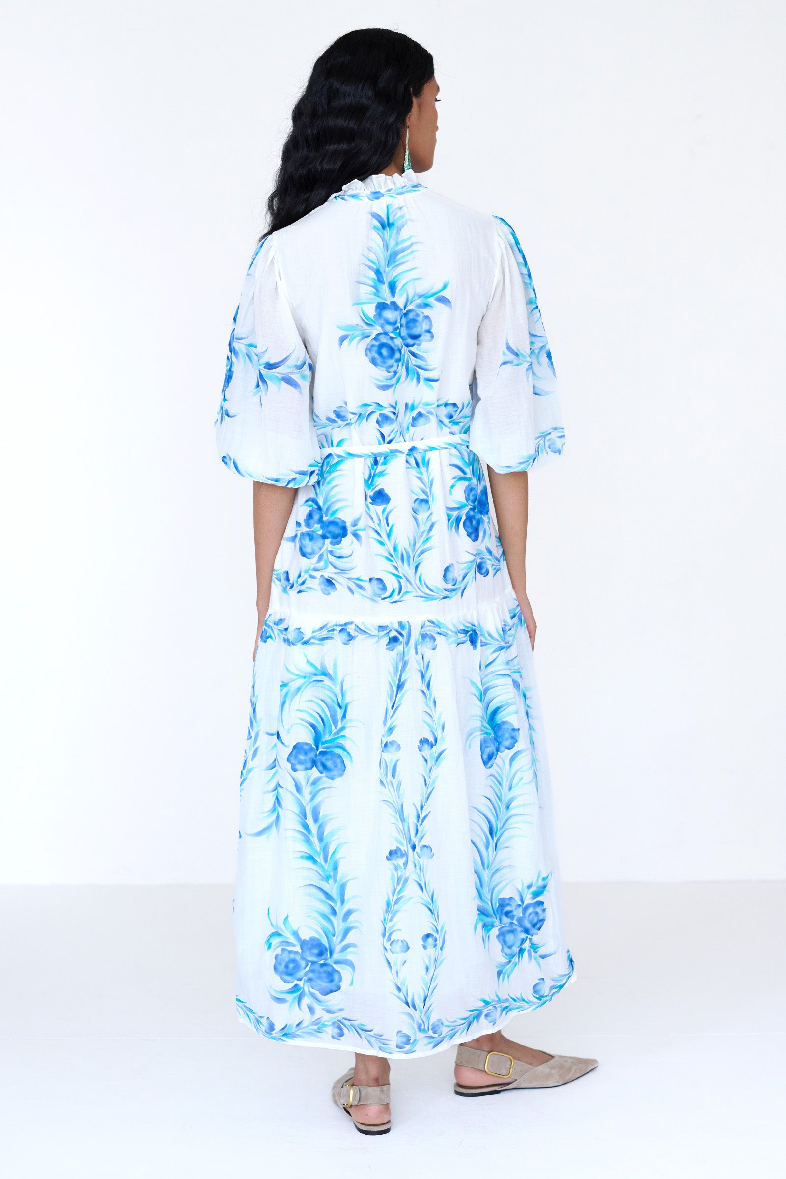 Back view of a woman standing wearing the Liza dress in cotton silk. It features hand-painted floral print, airy silhouette with fabric belt, tie and ruffled neckline.