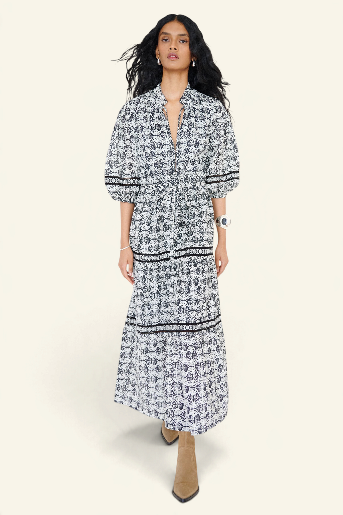 Front view of a woman walking wearing the Liza dress in Flutter print, featuring a loose silhouette, fabric belt, ruffled neckline with tie and bead details, Celtic-knot lace insets, and pockets.
