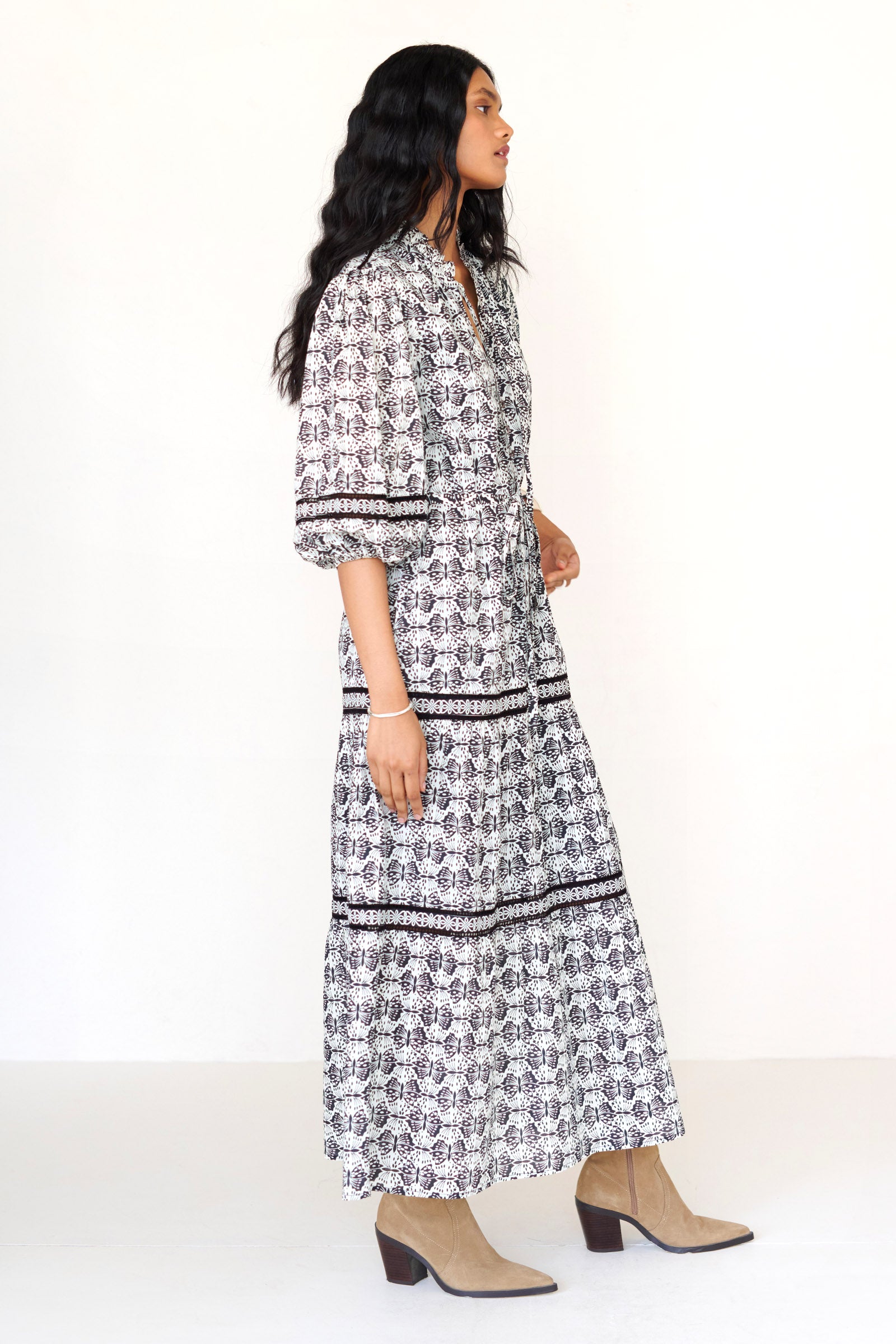 Side view of a woman standing wearing the Liza dress in Flutter print, featuring a loose silhouette, fabric belt, ruffled neckline with tie and bead details, Celtic-knot lace insets, and pockets.