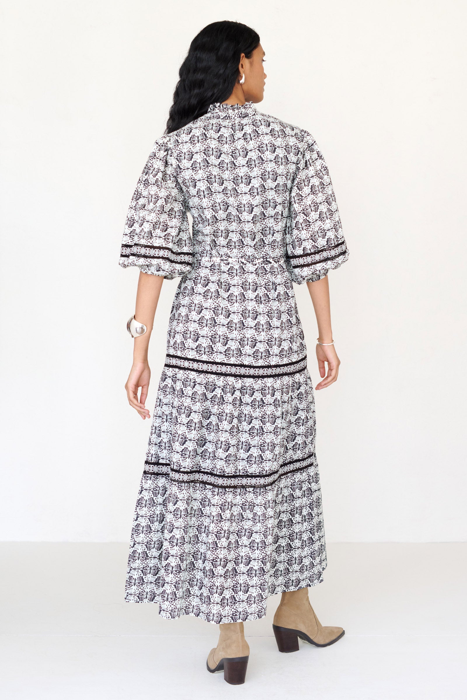 Back view of a woman standing wearing the Liza dress in Flutter print, featuring a loose silhouette, fabric belt, ruffled neckline with tie and bead details, Celtic-knot lace insets, and pockets.