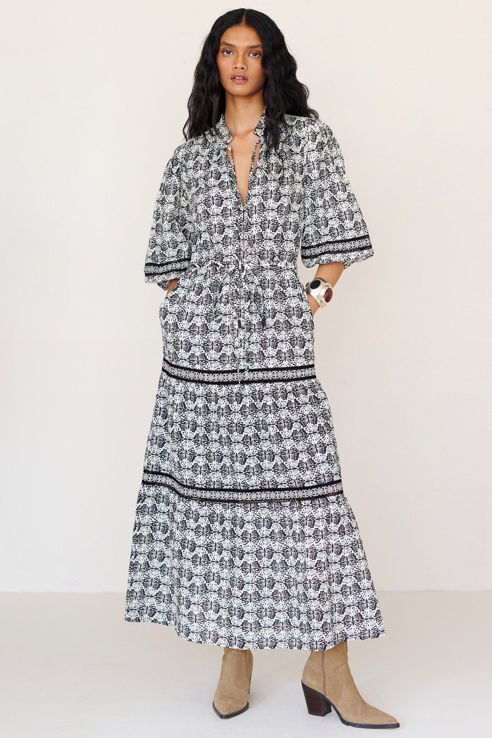 Front view of a woman standing with hands in pocket. She is wearing the Liza dress in Flutter print. It features a loose, airy silhouette, fabric belt, tie-neck with bead details, ruffled neckline, and side-seam pockets.