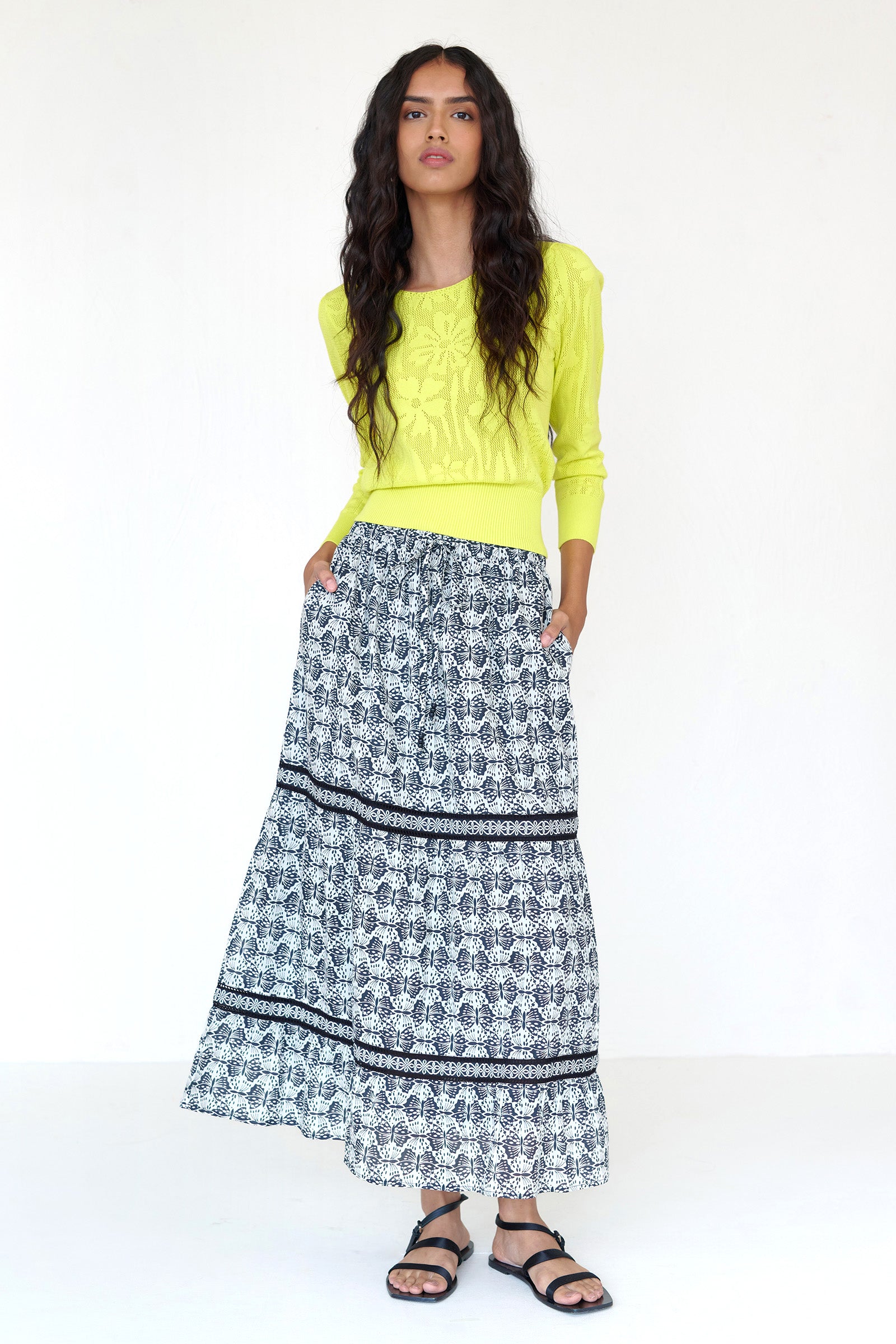 Front view of a woman standing. She wearing the Liza tiered skirt in Flutter print. It features an elastic drawstring waistband, Celtic-knot lace insets, and breezy cotton voile, styled with a neon yellow sweater.