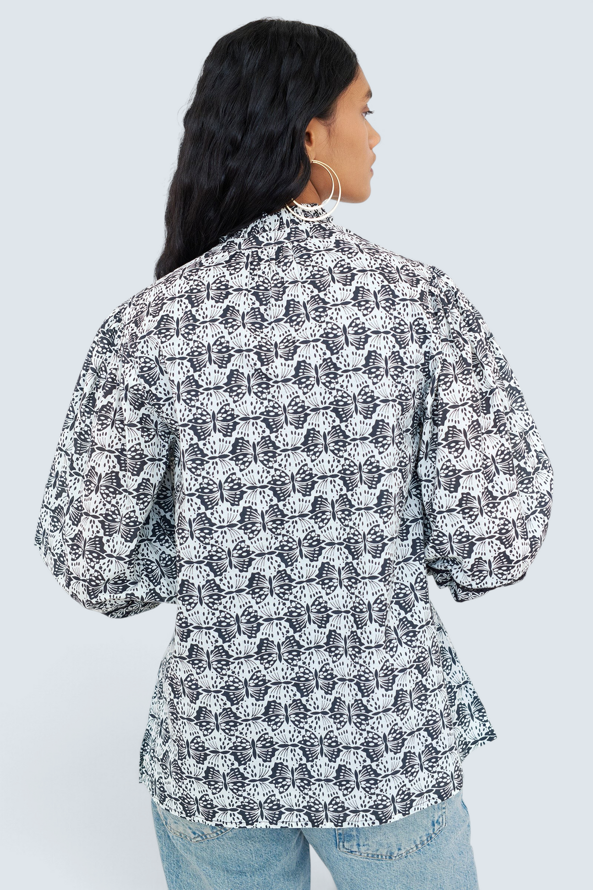 Back view of a woman standing wearing the Lorna top in Flutter print, featuring a ruffled V-neck with adjustable tie, Celtic-knot lace sleeve inserts, and a loose cotton voile silhouette styled with jeans