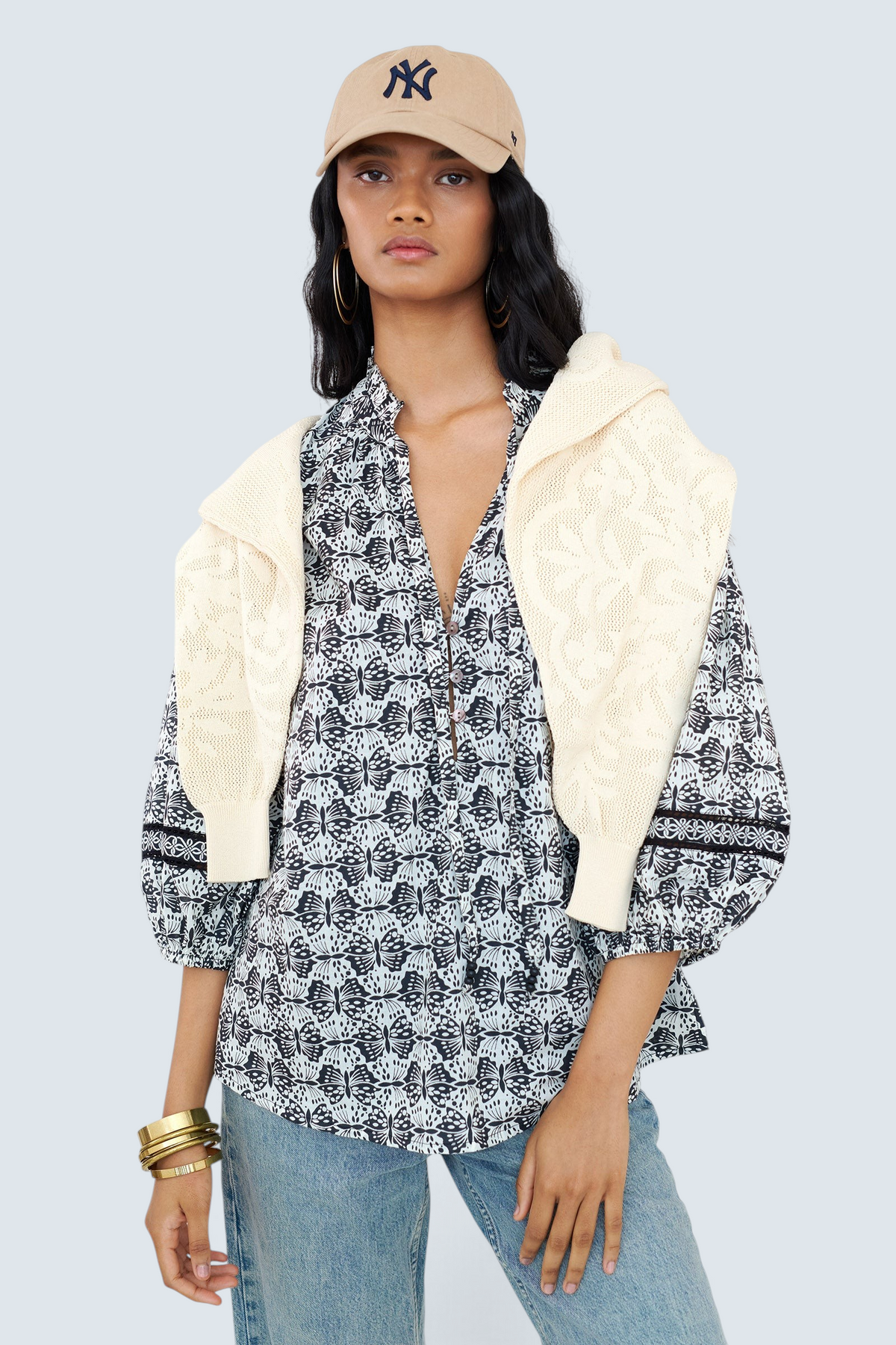 Front view of a woman standing wearing the Lorna top in Flutter print, featuring a ruffled V-neck with adjustable tie, Celtic-knot lace sleeve inserts, and a loose cotton voile silhouette styled with jeans, cap, layered with a sweater on shoulders.